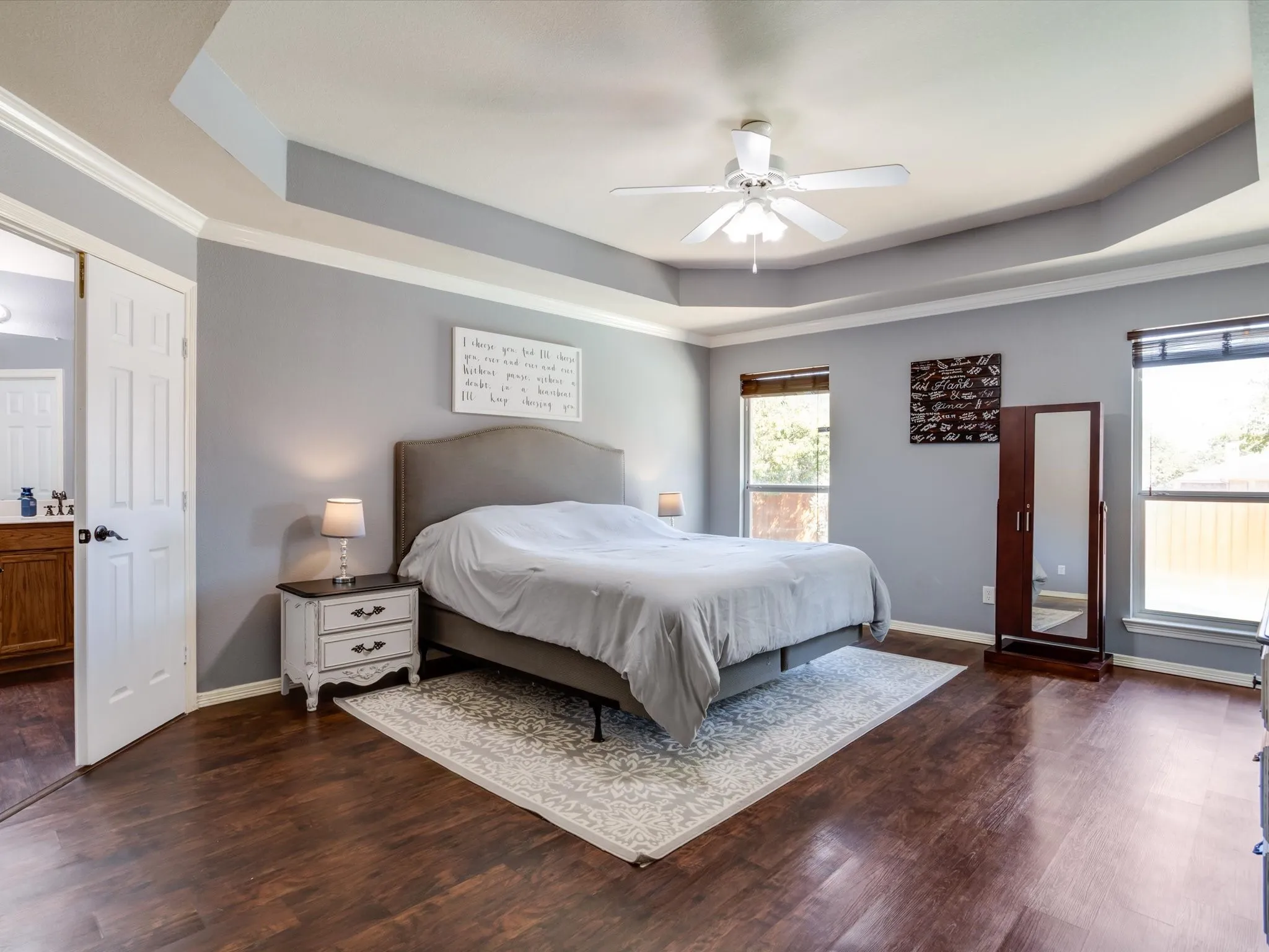 Tray ceiling with crown molding, ceiling fan, two windows, wood-finish flooring, and a paneled door