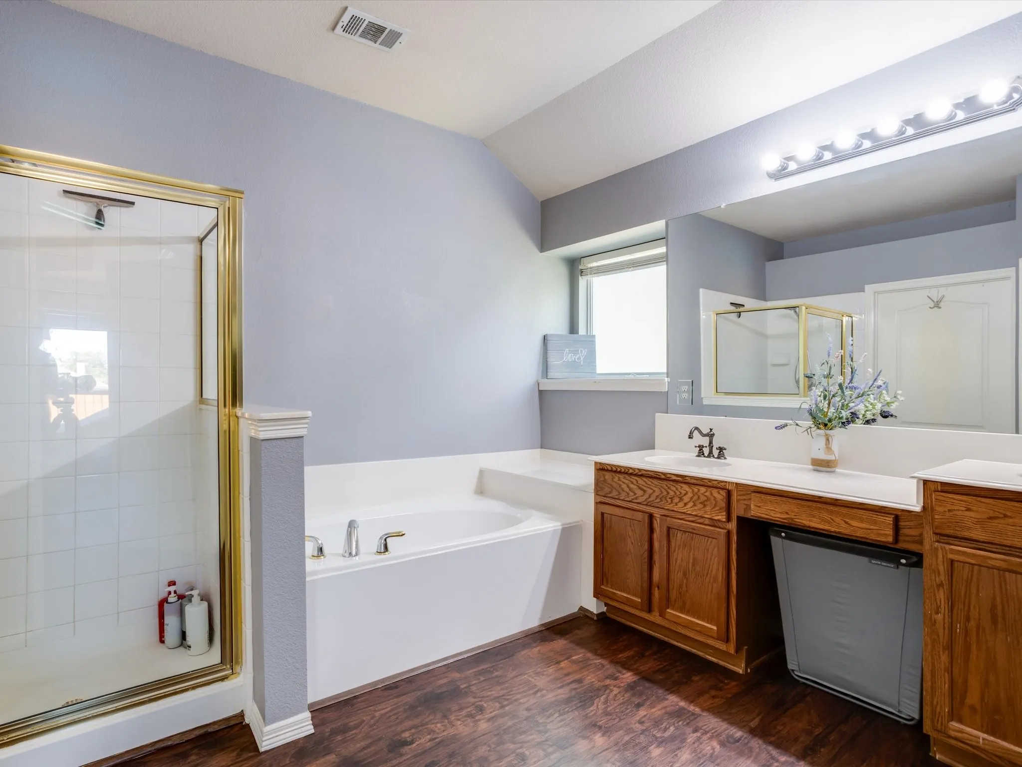Wood-finish flooring with a built-in tub, a gold-framed shower, and a vanity with wood cabinetry