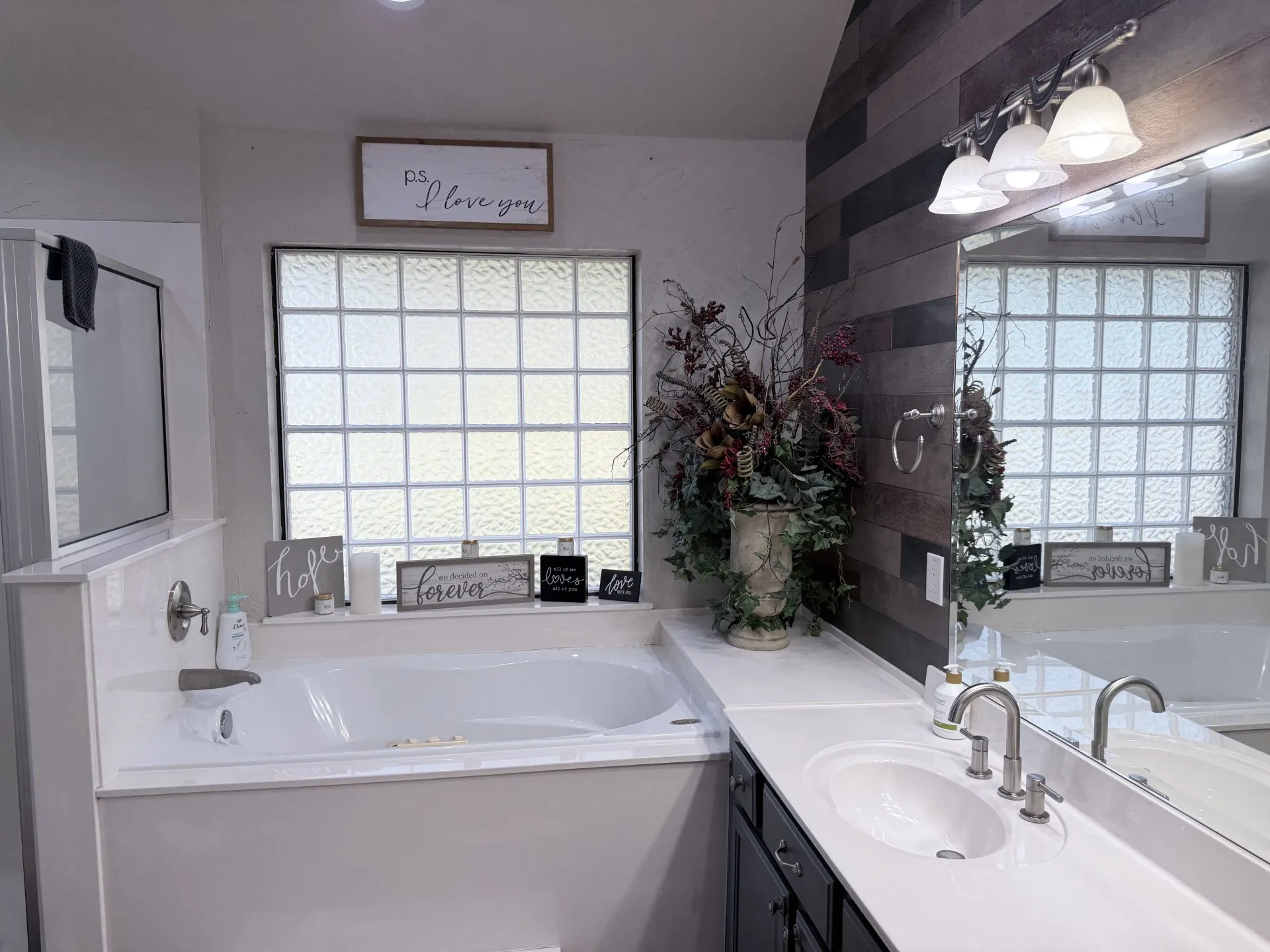 Built-in soaking tub, privacy glass block window, dual vanity with undermount sinks, wood-finish accent wall, and integrated vanity lighting