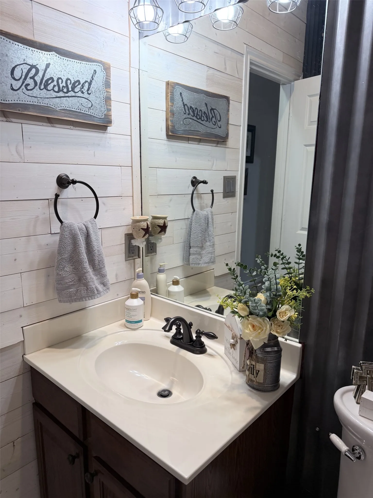 Shiplap-style wall paneling, vanity with an integrated sink, dark-finish vanity cabinet, dark-finish faucet, and overhead lighting with wire-frame shades