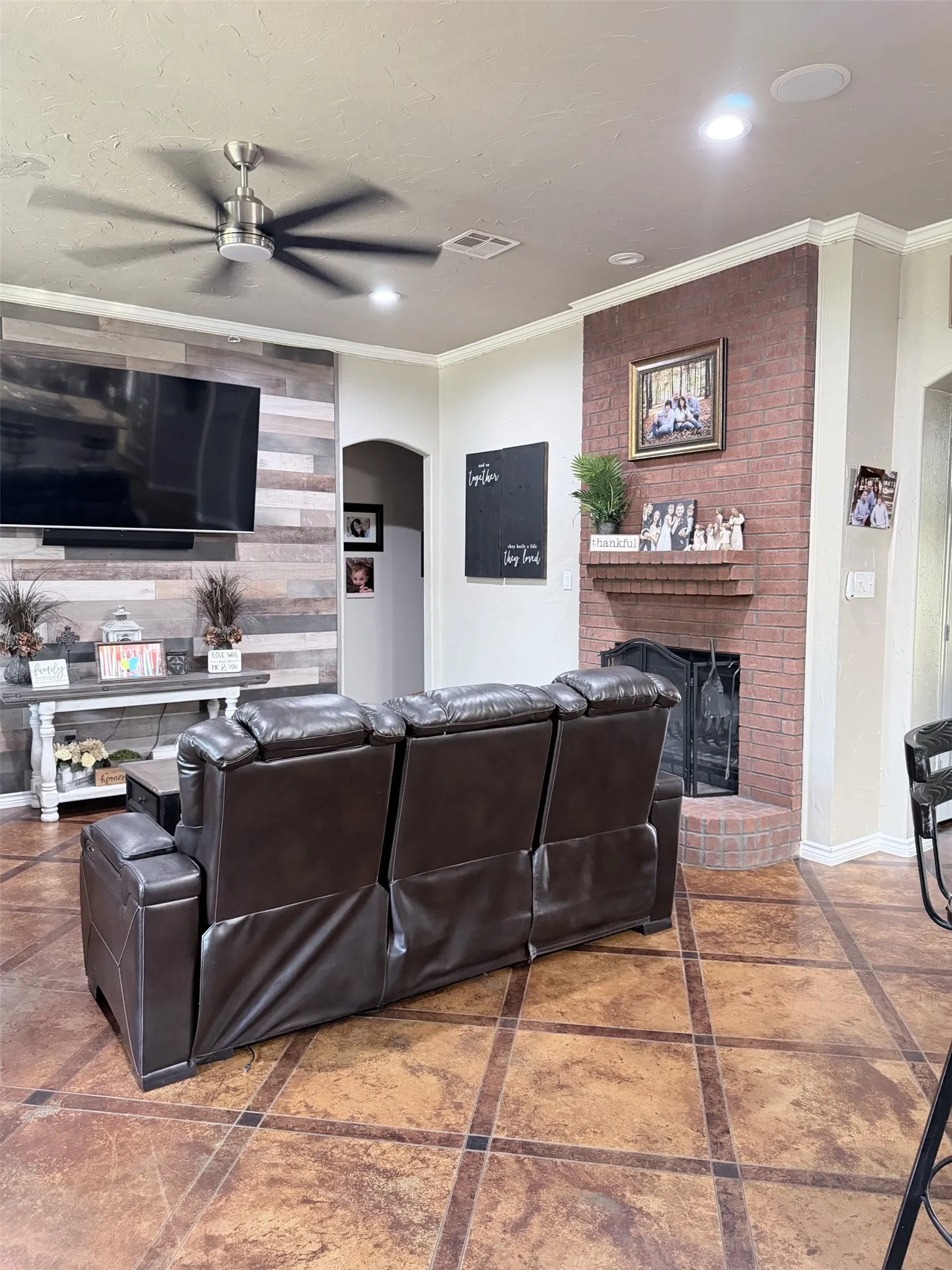 Recessed lighting, wood-plank accent wall, brick fireplace with mantel, ceiling fan, and patterned tile flooring