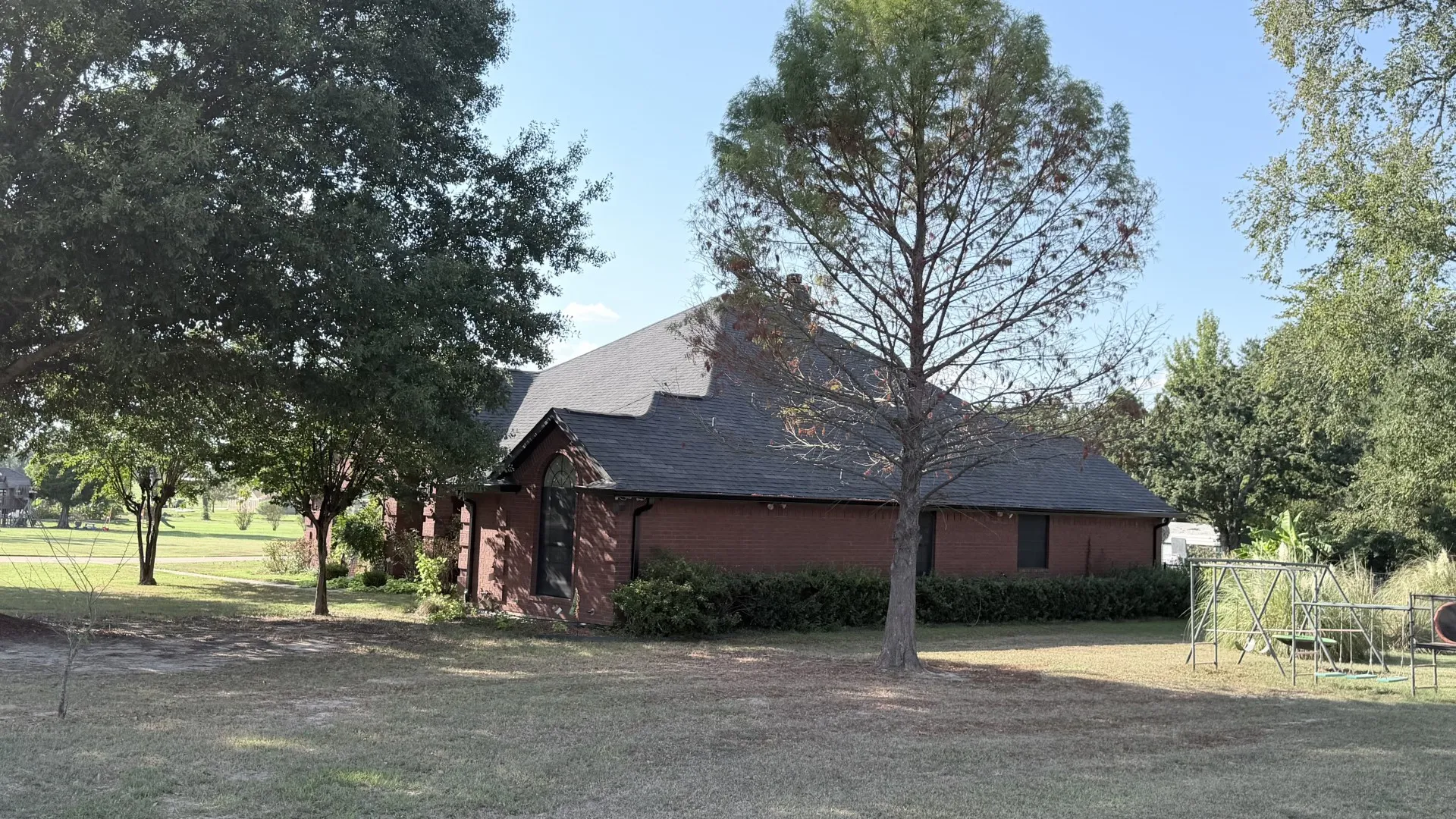 Brick exterior home featuring multiple mature trees and a sloped shingle roof