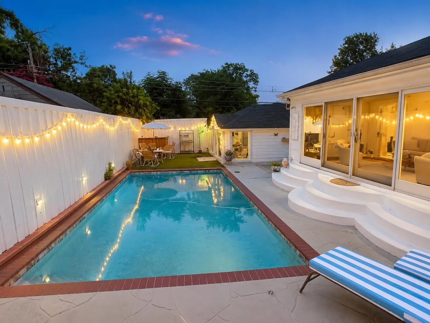 Rectangular swimming pool with brick coping, patio with stone-finish pavers, white privacy fencing, and multiple sliding glass doors