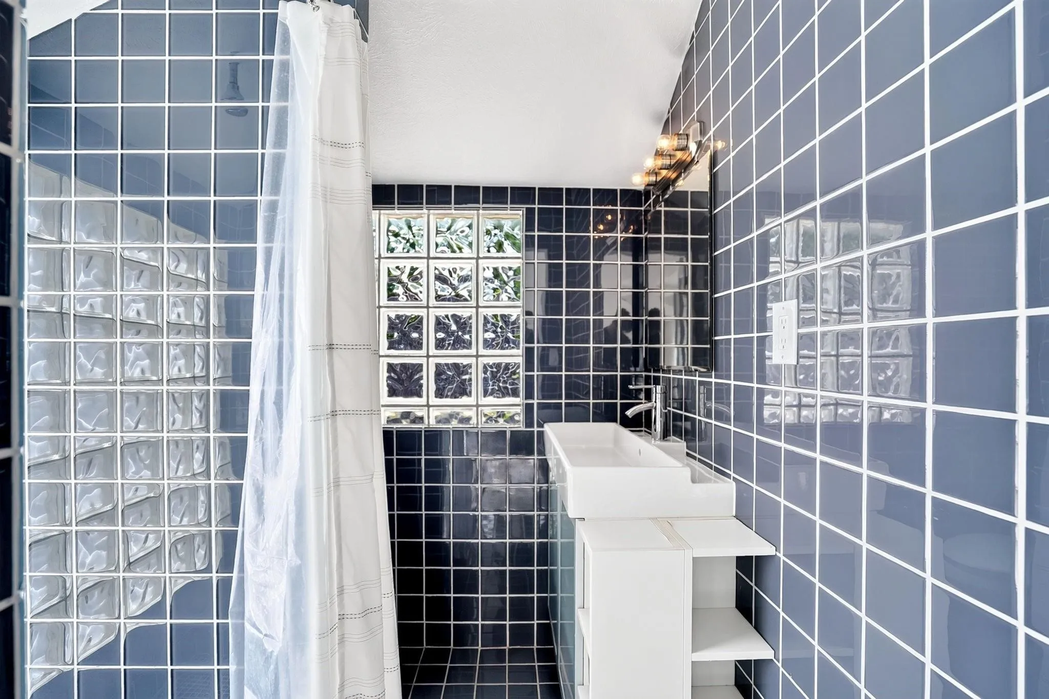 Bathroom featuring blue square wall tiles with white grout, glass block window, and a wall-mounted vanity with integrated sink