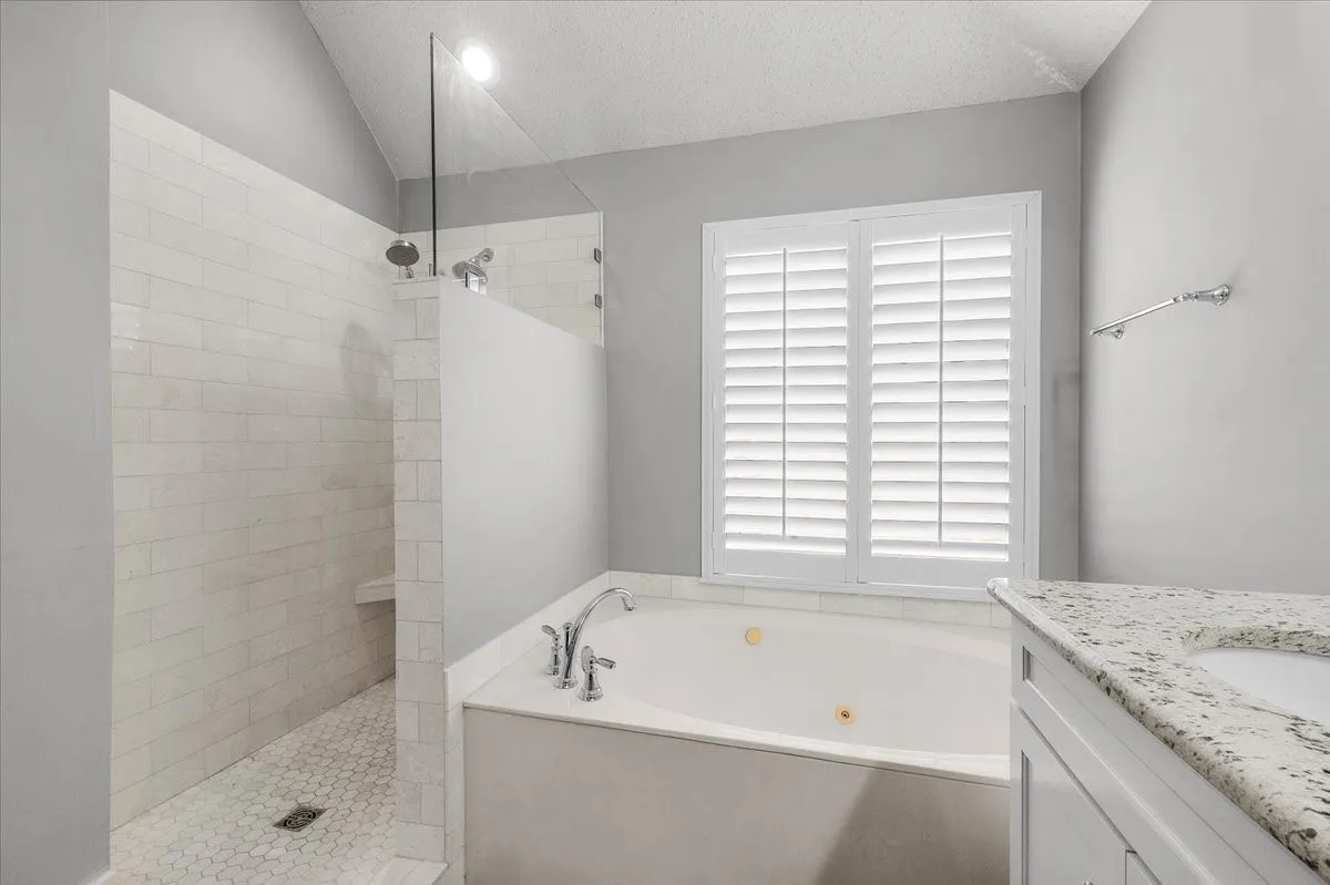 Vaulted ceiling, walk-in shower with bench and dual shower heads, jetted soaking tub, plantation shutters, and granite-finish vanity