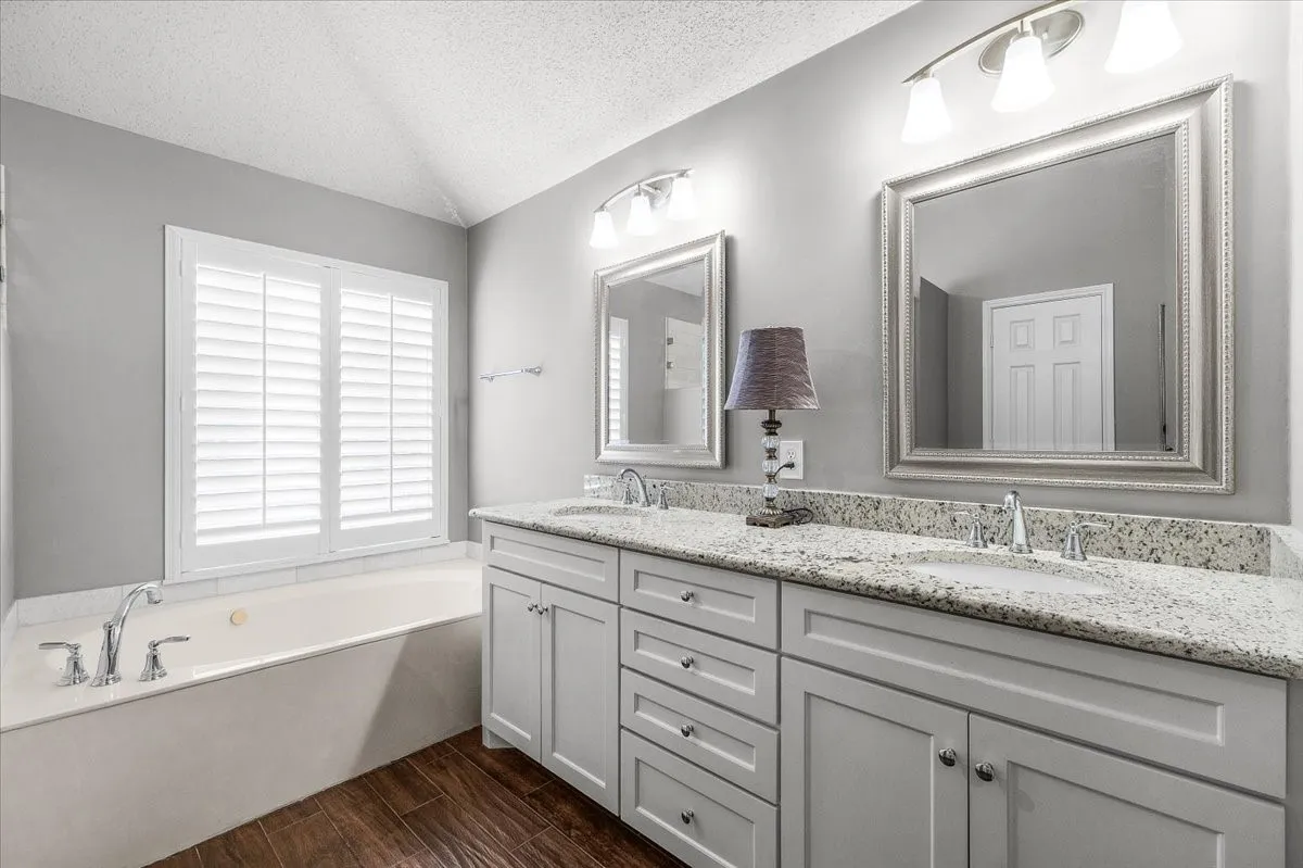 Dual vanity with granite countertops, wood-finish flooring, built-in soaking tub, and white plantation shutters