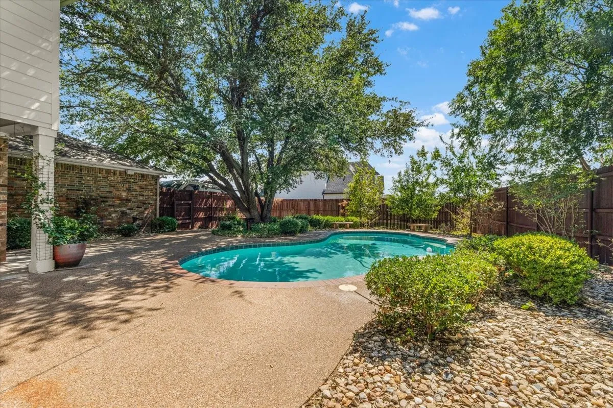 Curved swimming pool with surrounding aggregate concrete patio, mature trees providing shade, and a wood privacy fence
