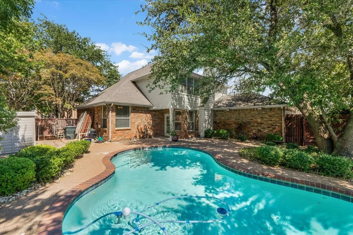 Curved swimming pool with brick coping, brick exterior with white siding, and mature trees