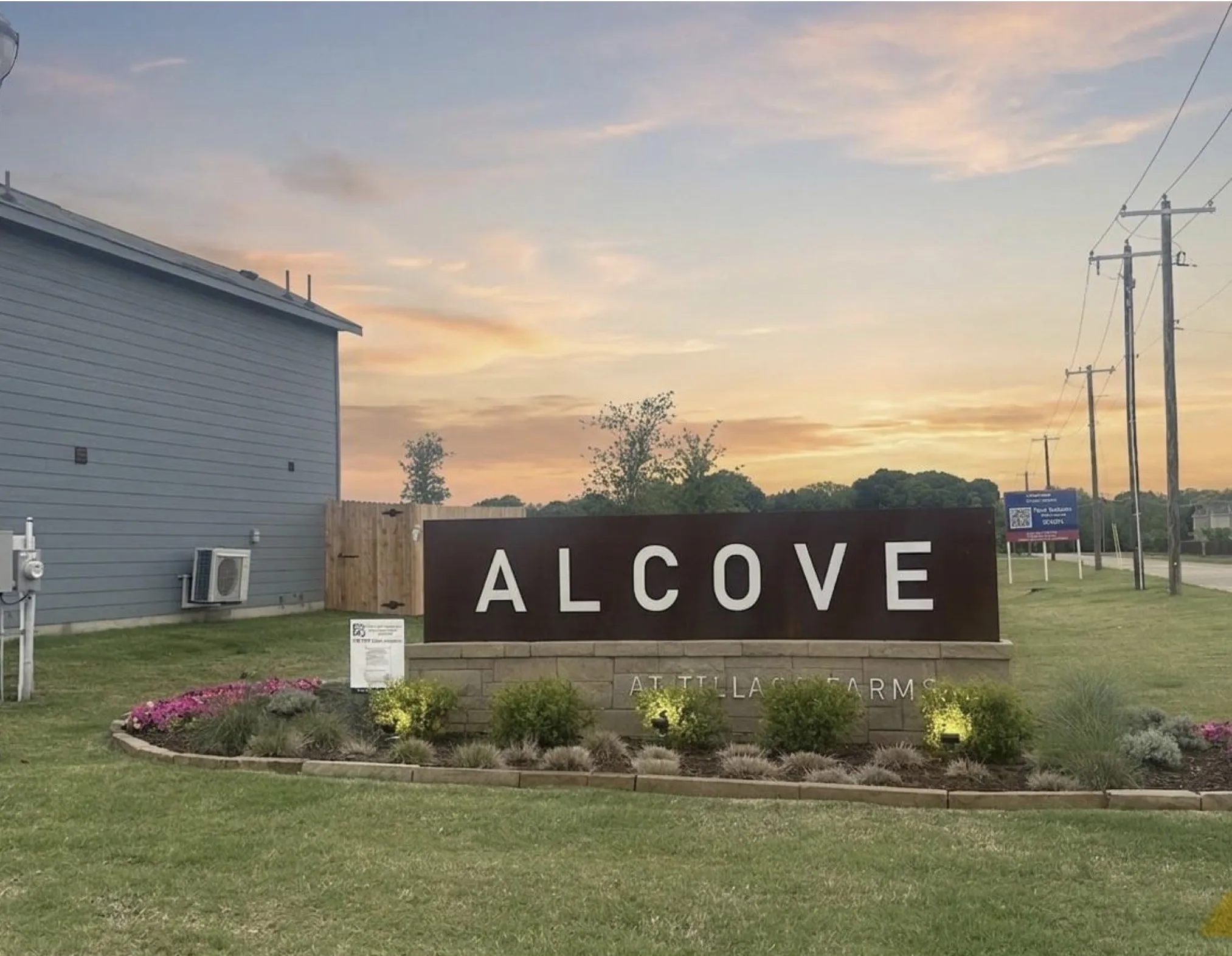 Community entrance signage featuring a stone base and metal lettering, surrounded by manicured landscaping with flowering plants and ground lighting
