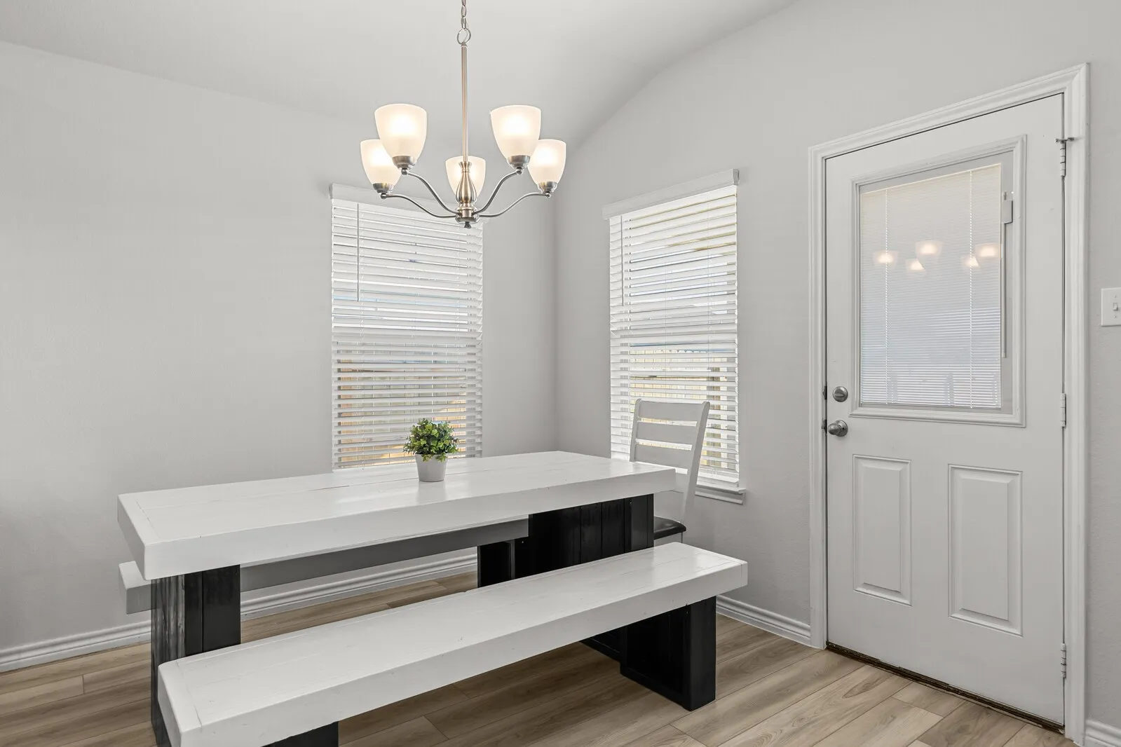 Vaulted ceiling with a five-light chandelier, two windows with blinds, wood-finish flooring, and a white paneled door with a privacy glass insert