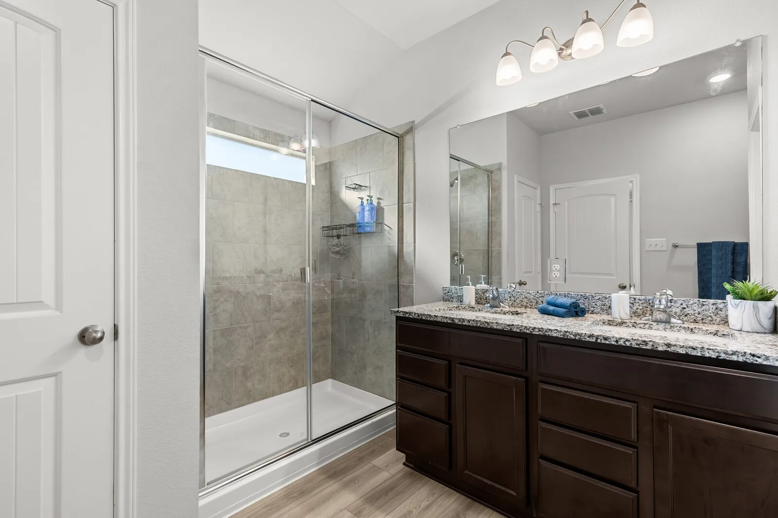 Spacious bathroom featuring a dual vanity with a patterned countertop, dark wood-finish cabinetry, a large frameless mirror, and a walk-in shower with tile surround