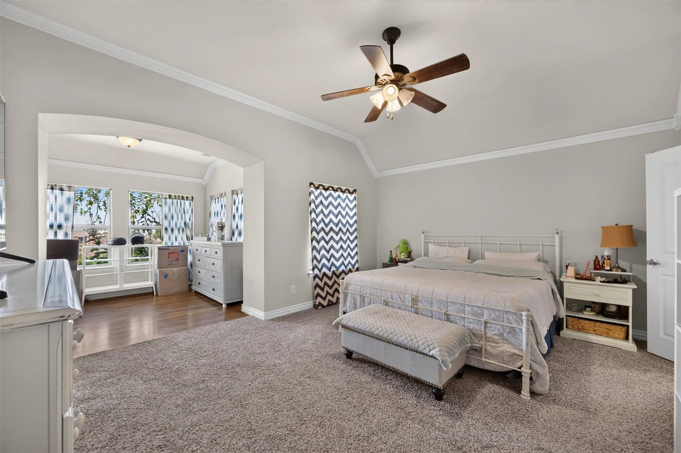 Vaulted ceiling with crown molding, ceiling fan with integrated lighting, arched entryway, wood-finish flooring, and multiple windows