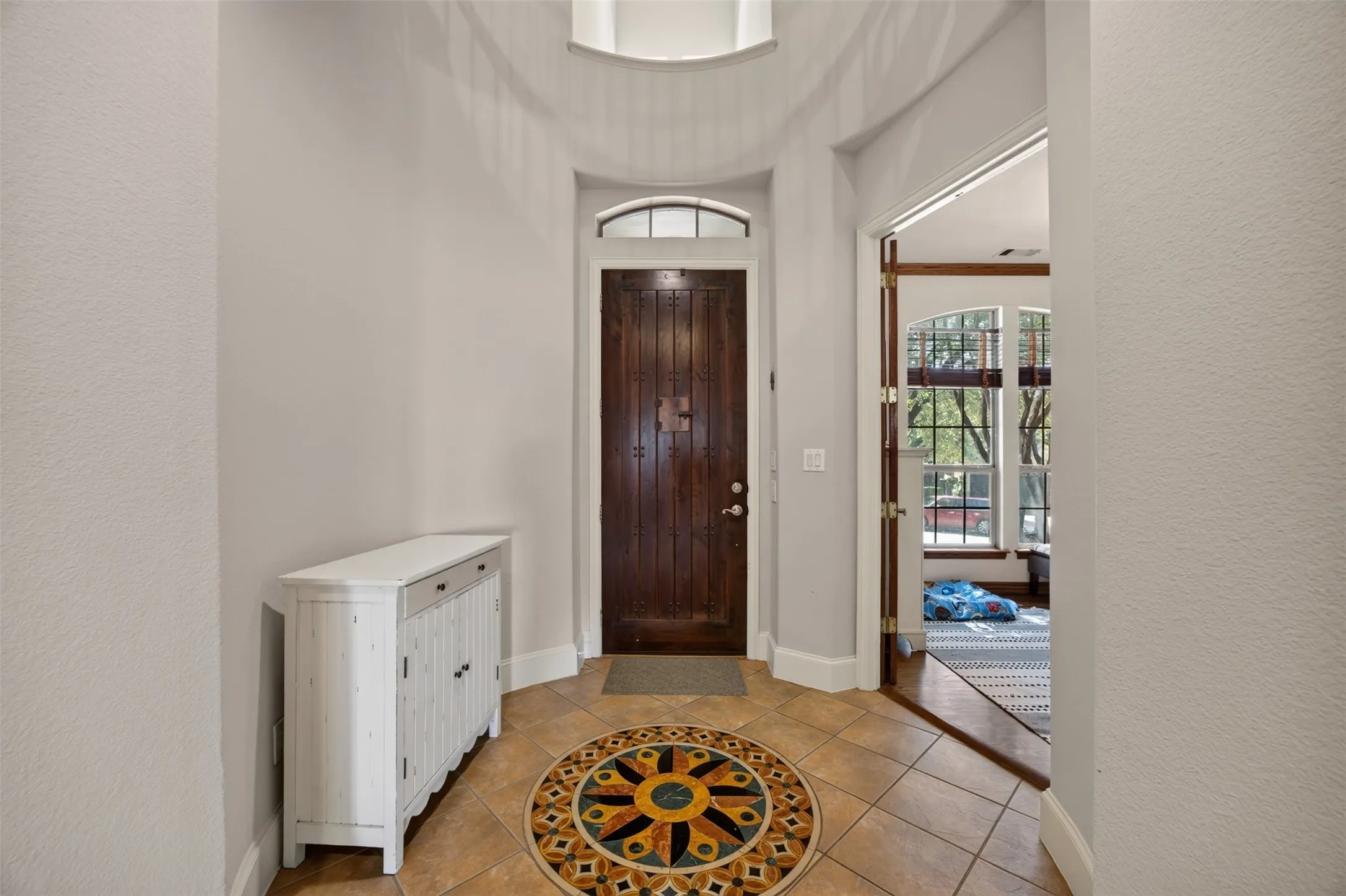 Entryway featuring a wood plank door with an arched transom, tile flooring with an inlaid mosaic medallion, and an arched doorframe leading to an interior space with large windows