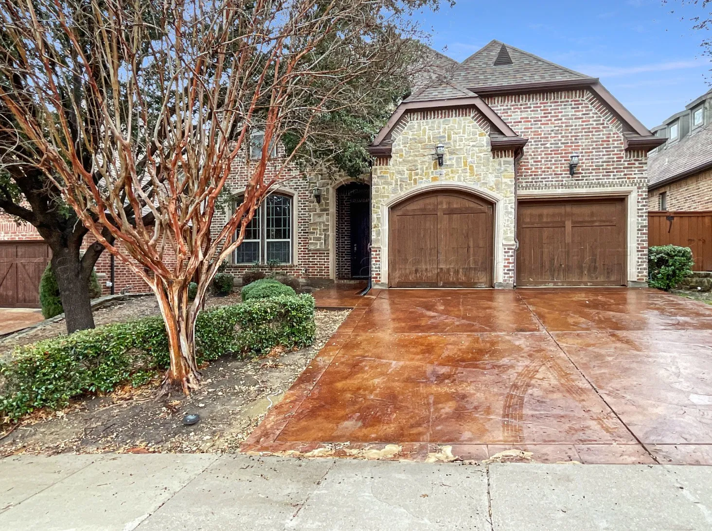Brick and stone facade featuring arched garage doors, decorative outdoor lighting, a patterned concrete driveway, and established landscaping