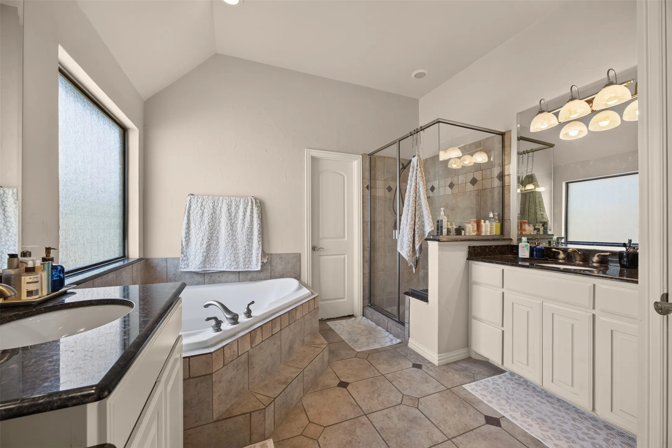 Soaking tub with tiled surround, walk-in shower with glass enclosure, dual vanity with dark countertops, and tile flooring