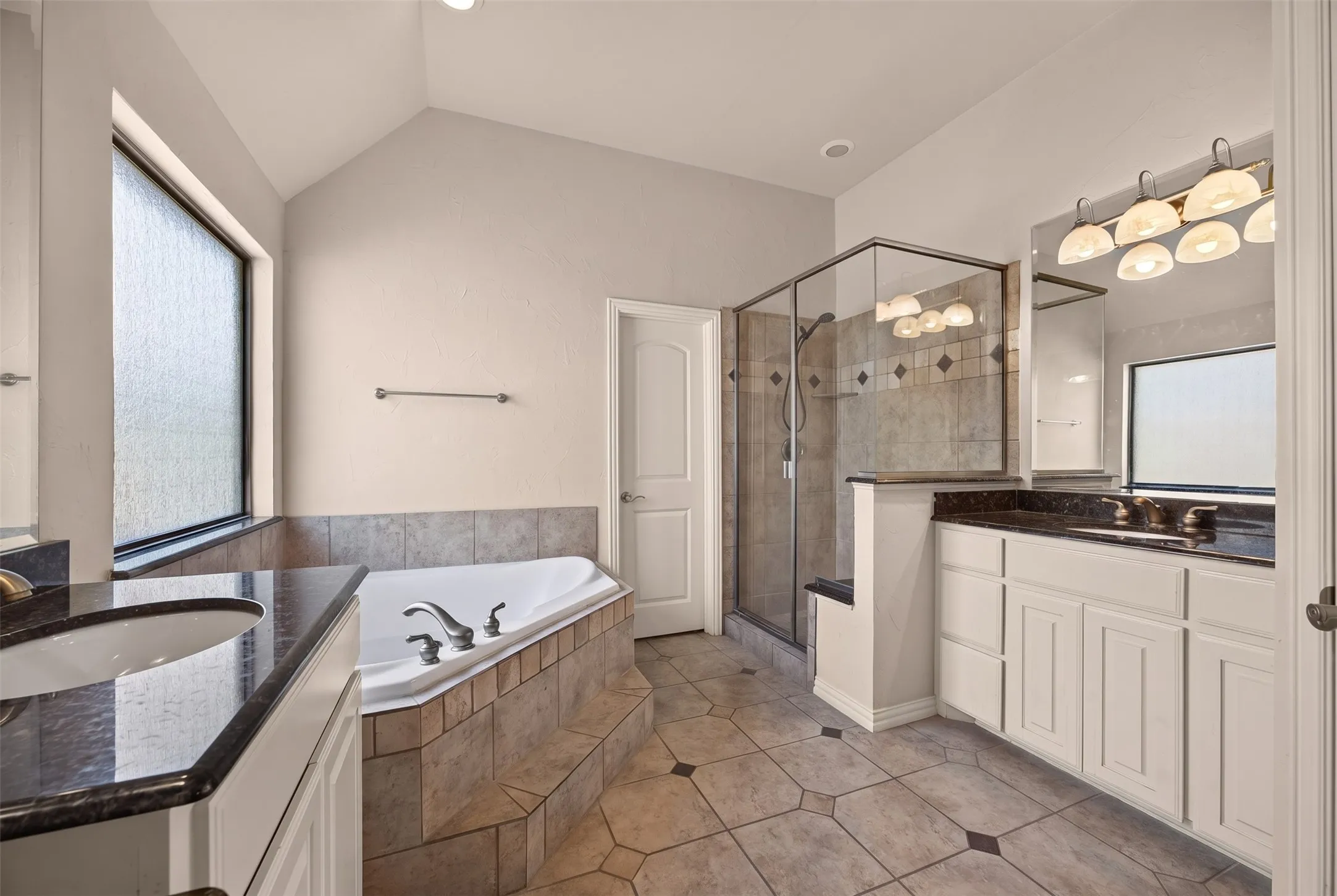 Soaking tub with tile surround, walk-in shower with glass enclosure, dual vanity with undermount sinks, dark countertops, and tile flooring