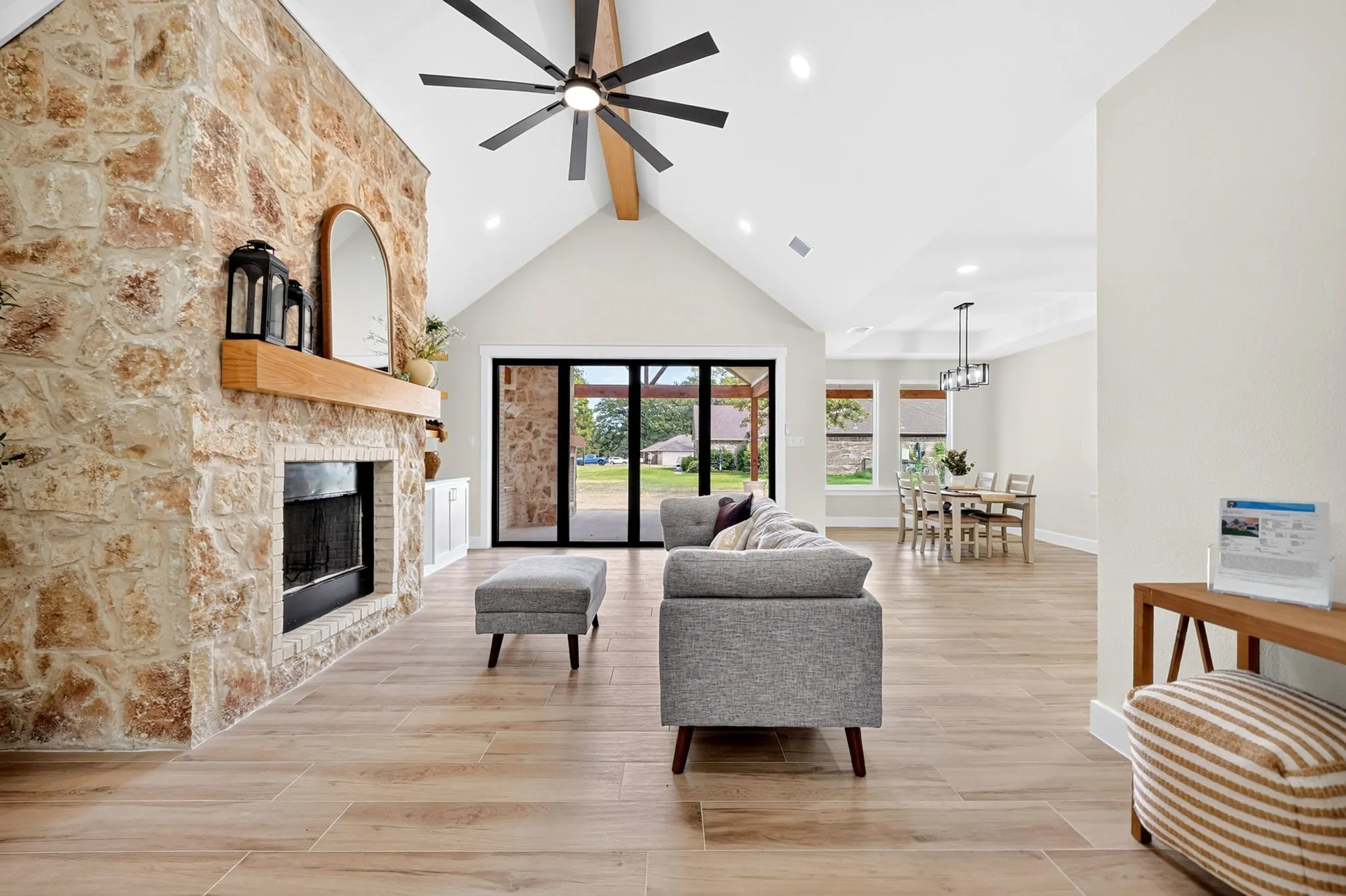Vaulted ceiling with exposed beam, stone fireplace with wood mantel, wood-finish flooring, black-framed sliding glass doors, and recessed lighting