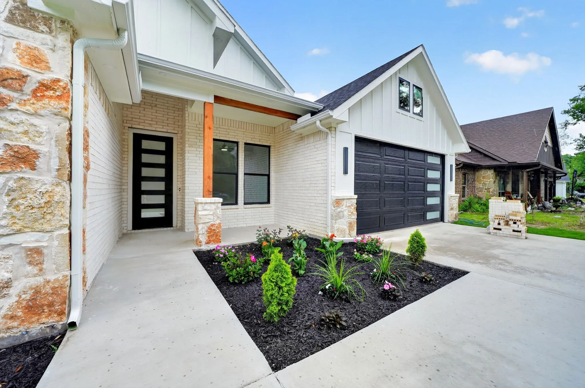 White brick exterior with stone accents, a modern black entry door, a dark garage door with horizontal windows, and a landscaped front planter