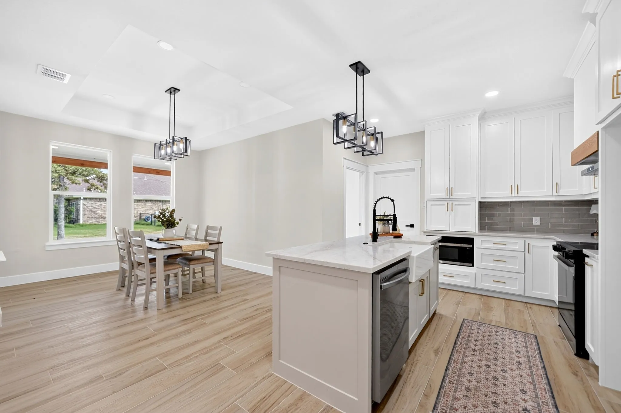 Light wood-finish flooring throughout, white shaker-style cabinetry, kitchen island with a farmhouse sink, stainless steel appliances, and a tray ceiling