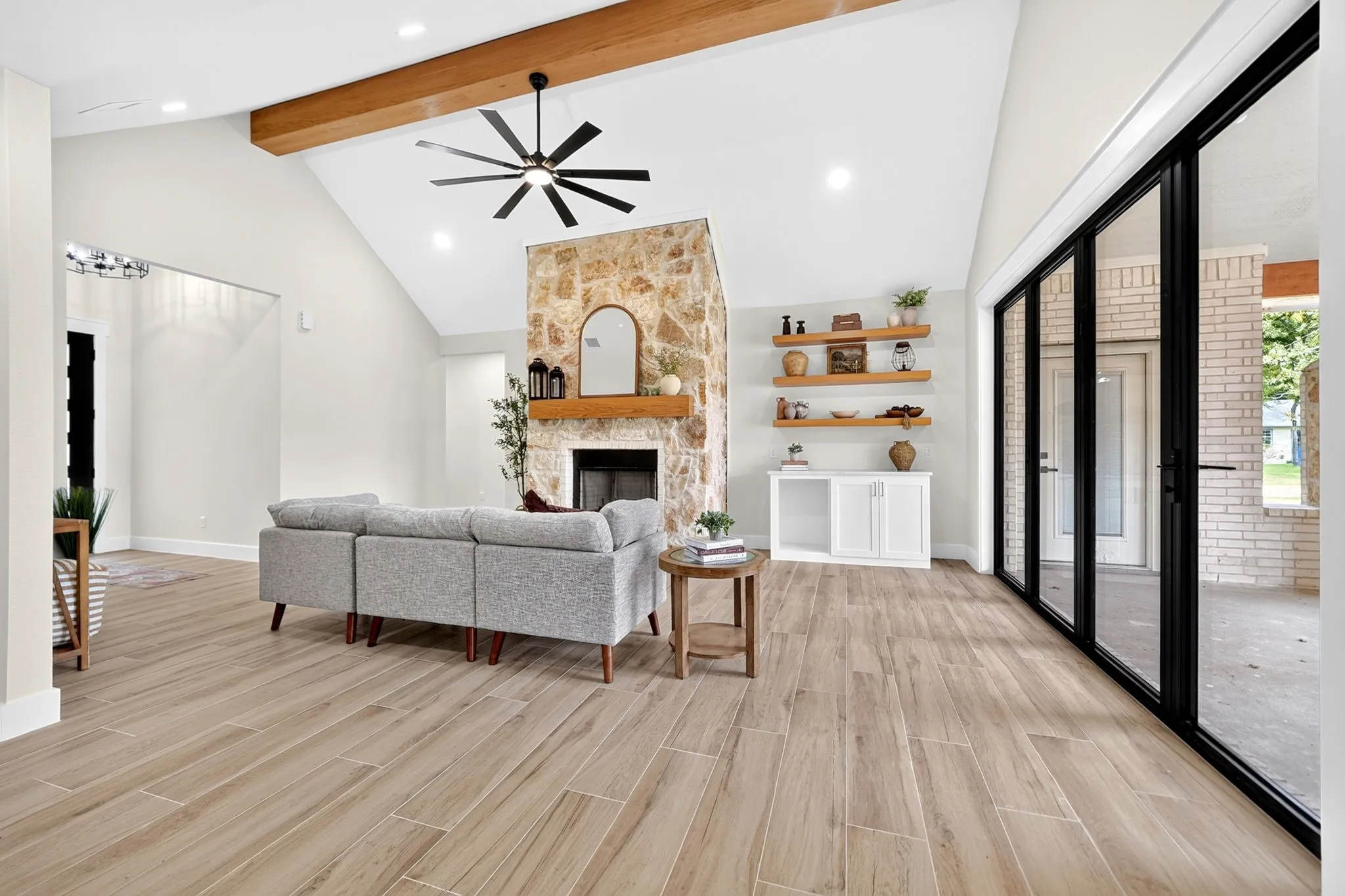 Vaulted ceiling with exposed wood beam, natural stone fireplace, wood-finish flooring, built-in shelving, and multi-panel sliding glass doors