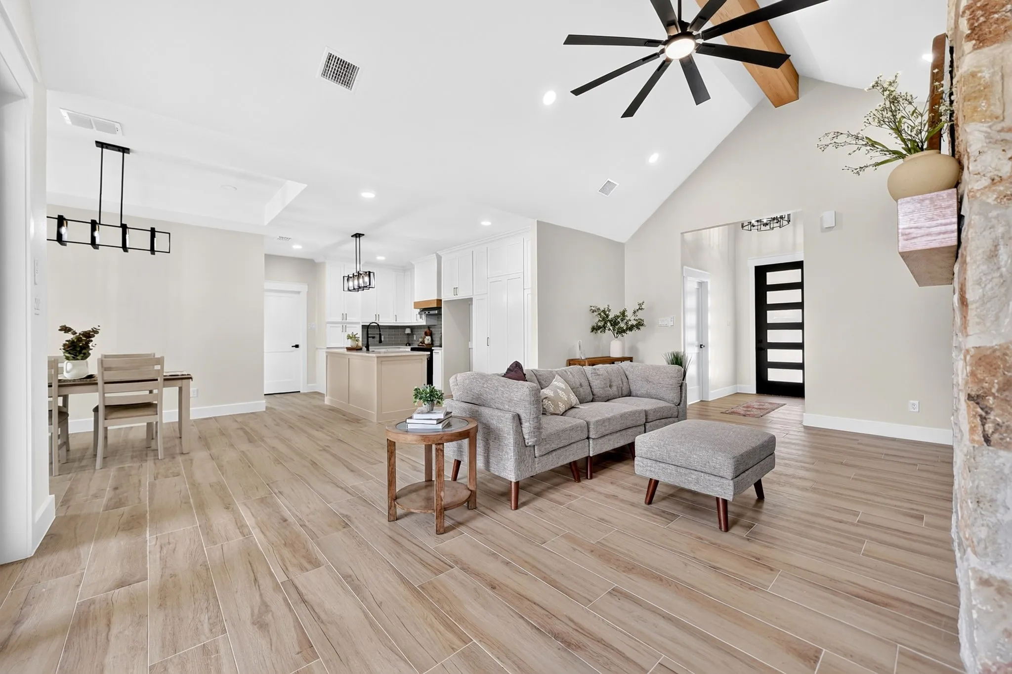 Vaulted ceiling with exposed wood beam, wood-finish flooring, stone accent wall, tray ceiling, and a contemporary linear chandelier