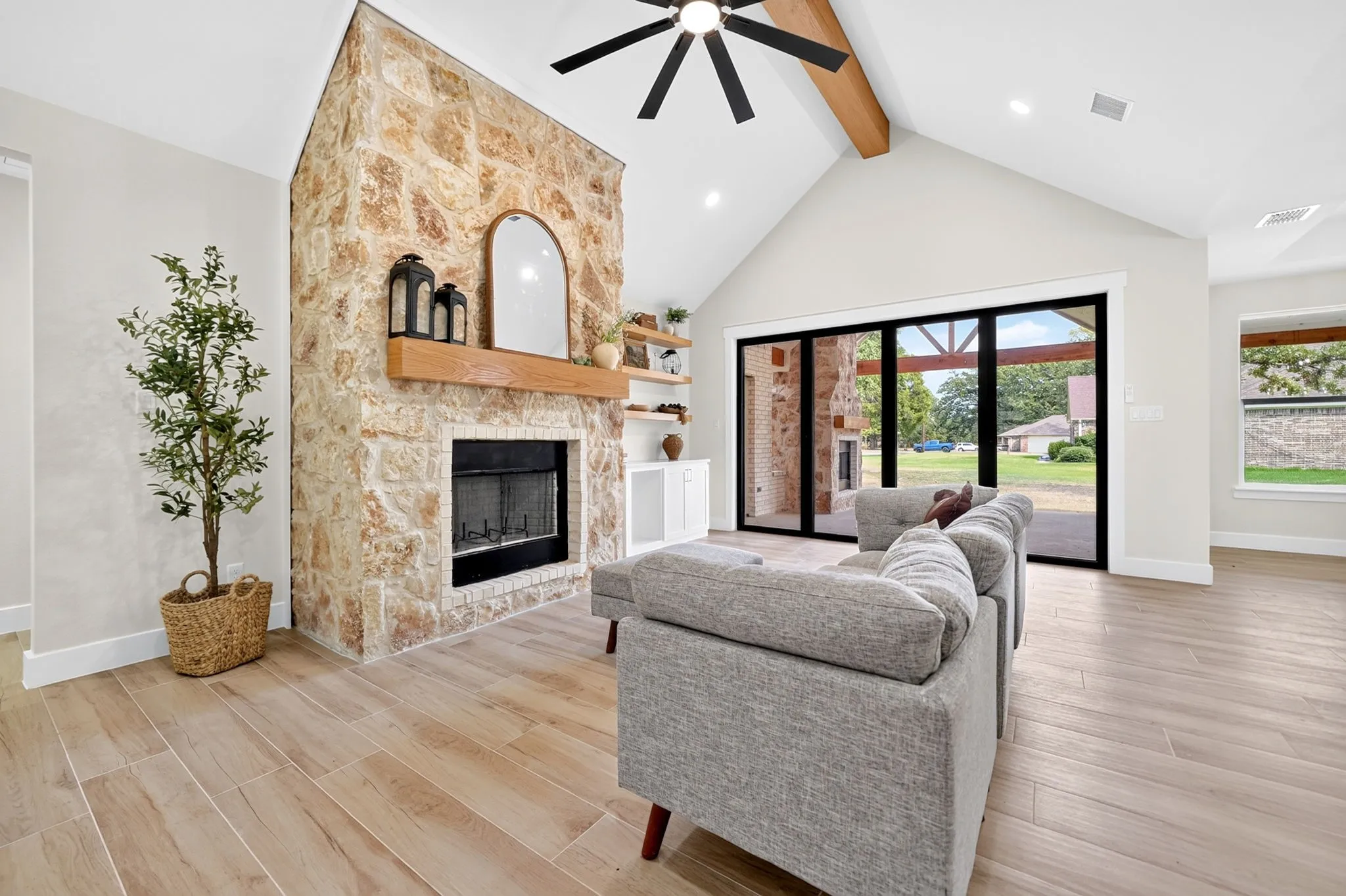 Vaulted ceiling with exposed beam, stone-clad fireplace with wood mantel, wood-finish flooring, and large sliding glass doors