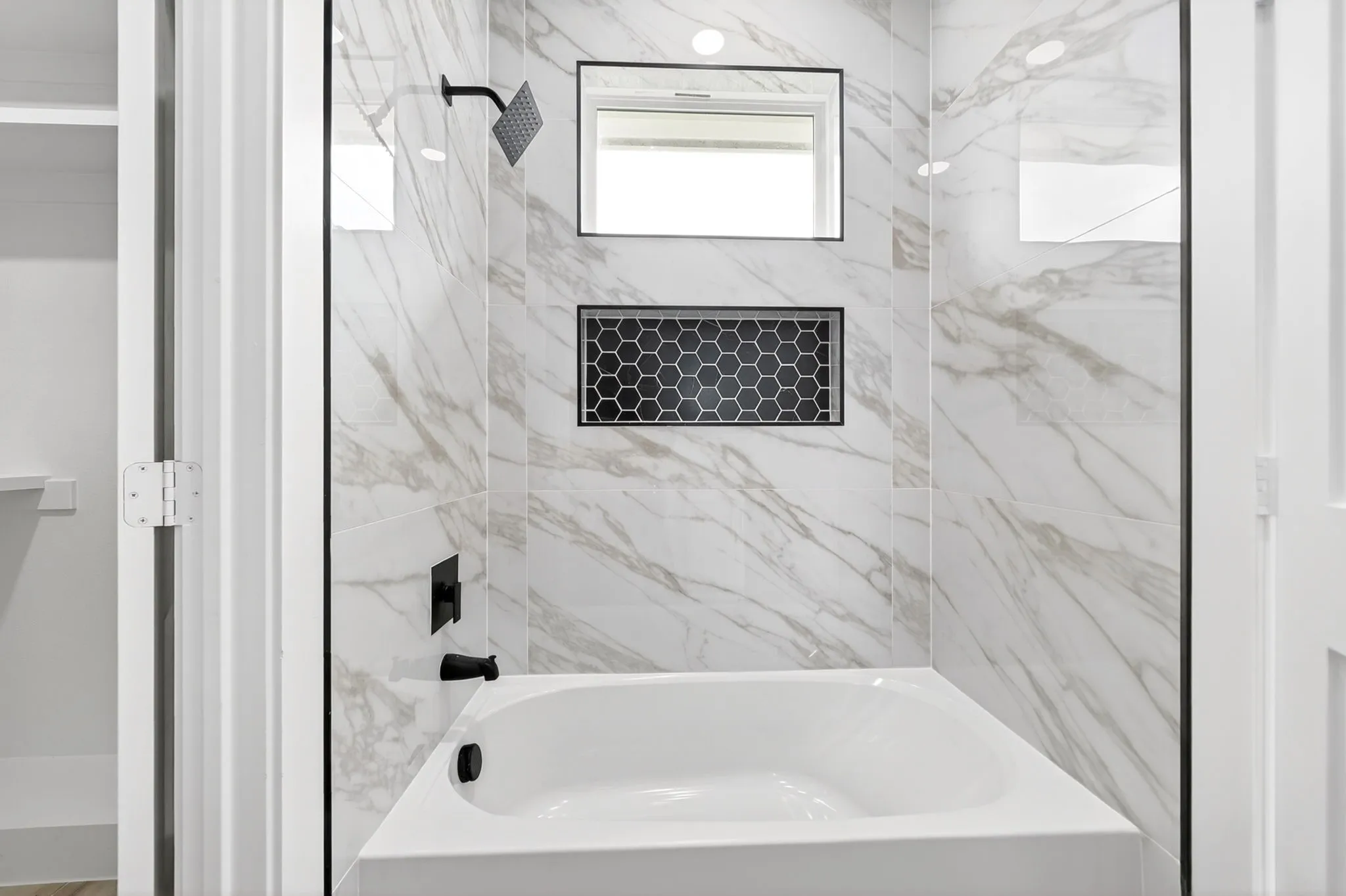 Bathtub with white porcelain finish, matte black fixtures, marble-look tiled surround, recessed shower niche with black hexagonal tile, and integrated window