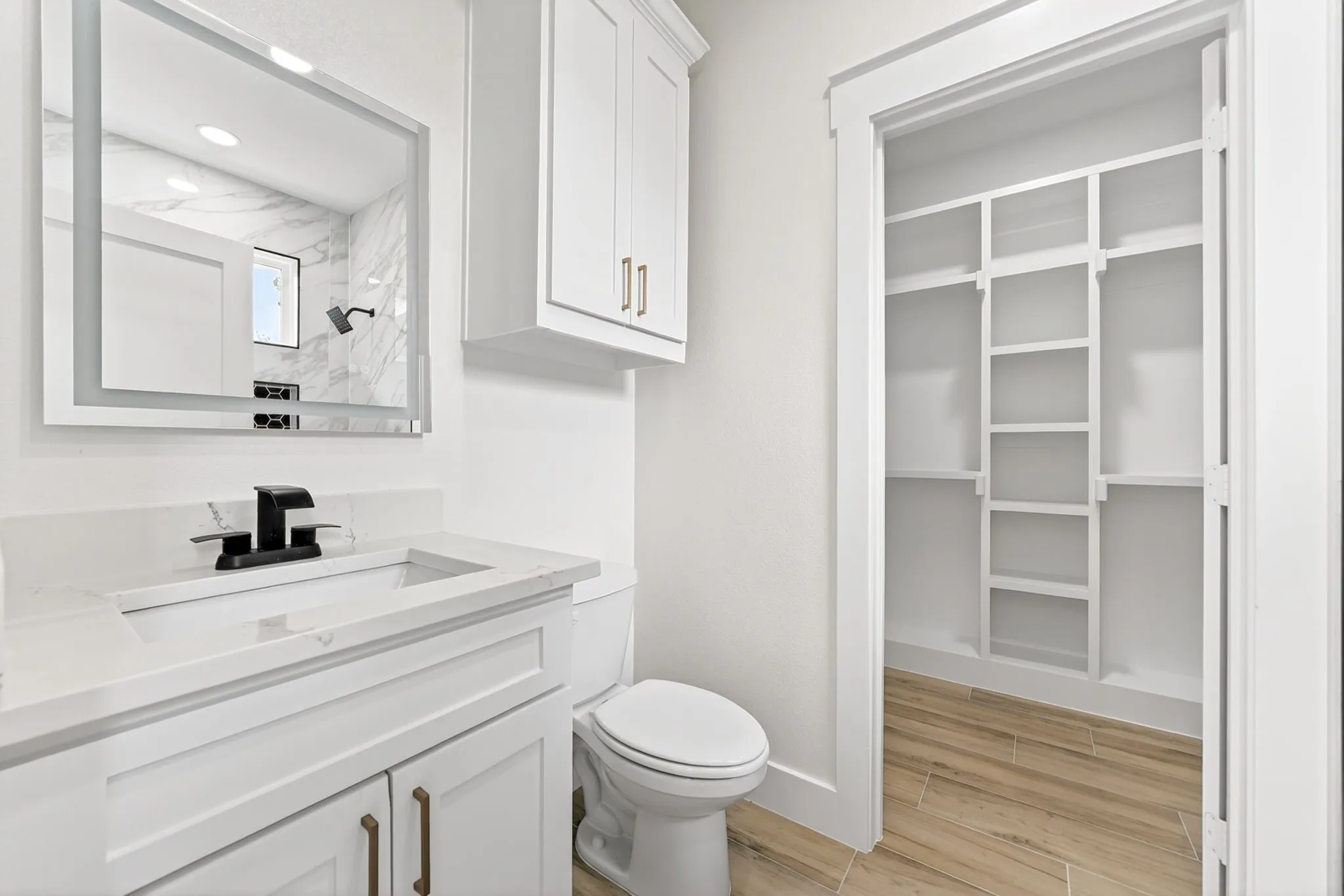 Bathroom featuring a single vanity with a white countertop and integrated sink, a wall-mounted lighted mirror, a white toilet, and wood-finish flooring