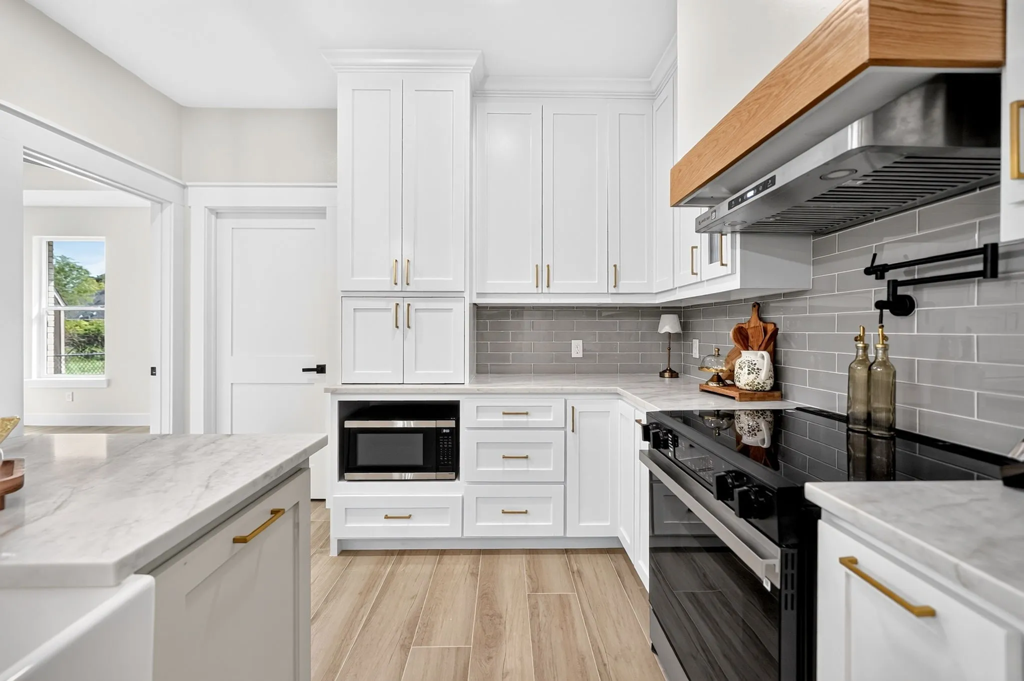 White shaker cabinetry with gold-finish hardware, light-toned wood-finish flooring, white countertops, gray subway tile backsplash, and a wooden range hood
