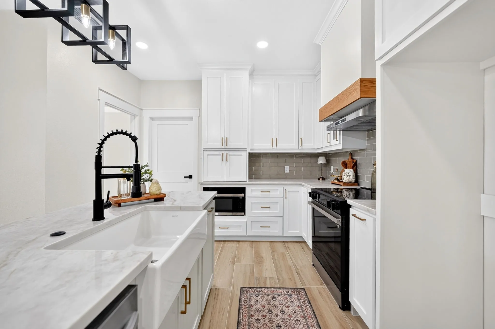 White shaker cabinetry, light-toned wood-finish flooring, white farmhouse sink, black coil spring faucet, and a subway tile backsplash