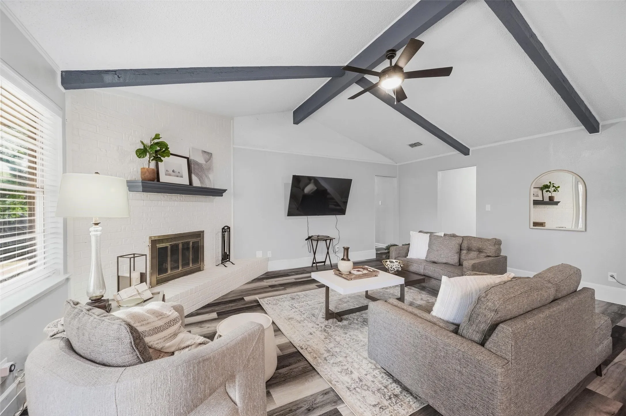 Vaulted ceiling with exposed beams, wood-finish flooring, white brick fireplace with mantel, ceiling fan, and large windows with blinds