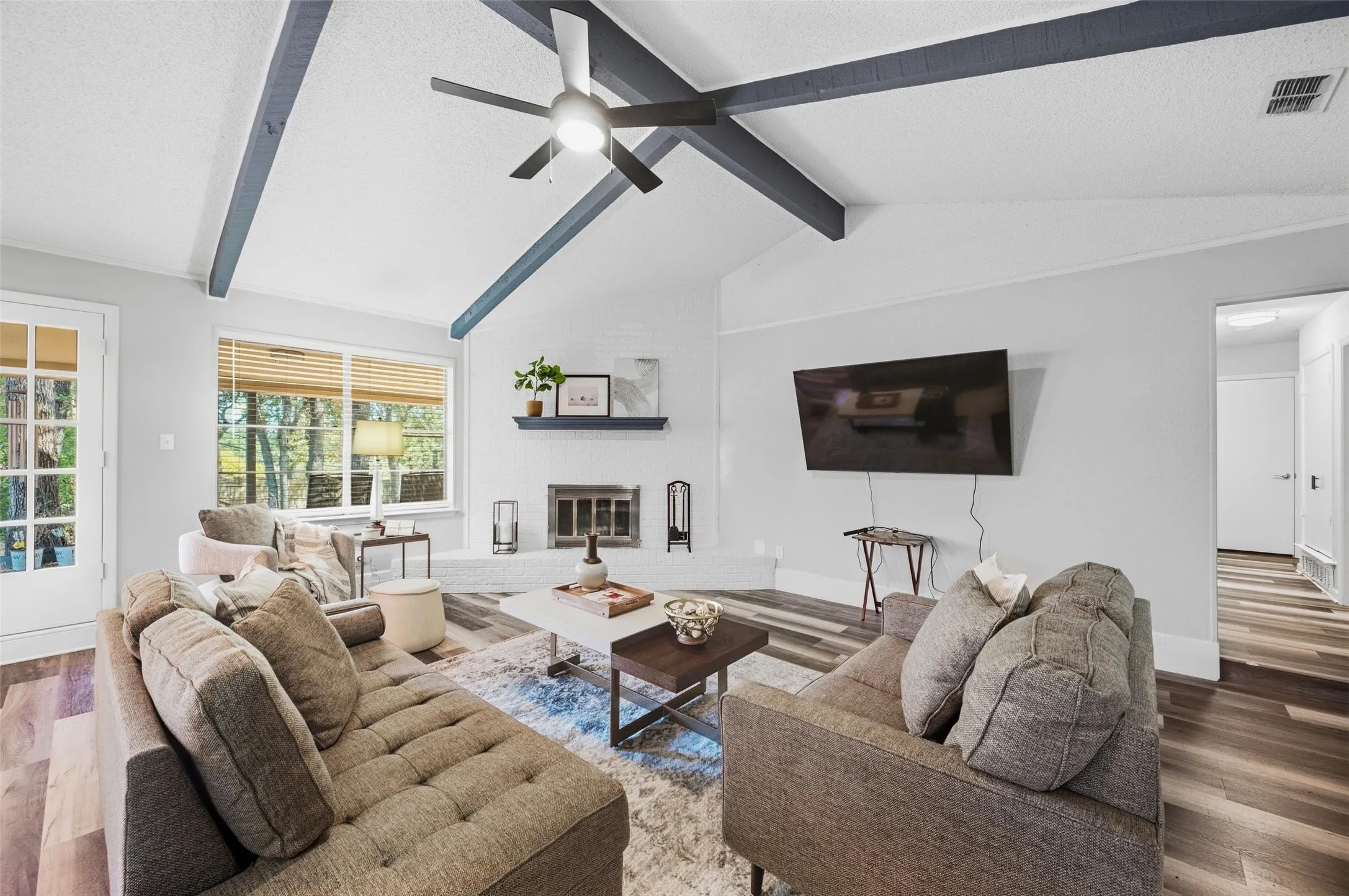 Vaulted ceiling with exposed beams, wood-finish flooring, white brick fireplace, large windows with natural light, and a white French door