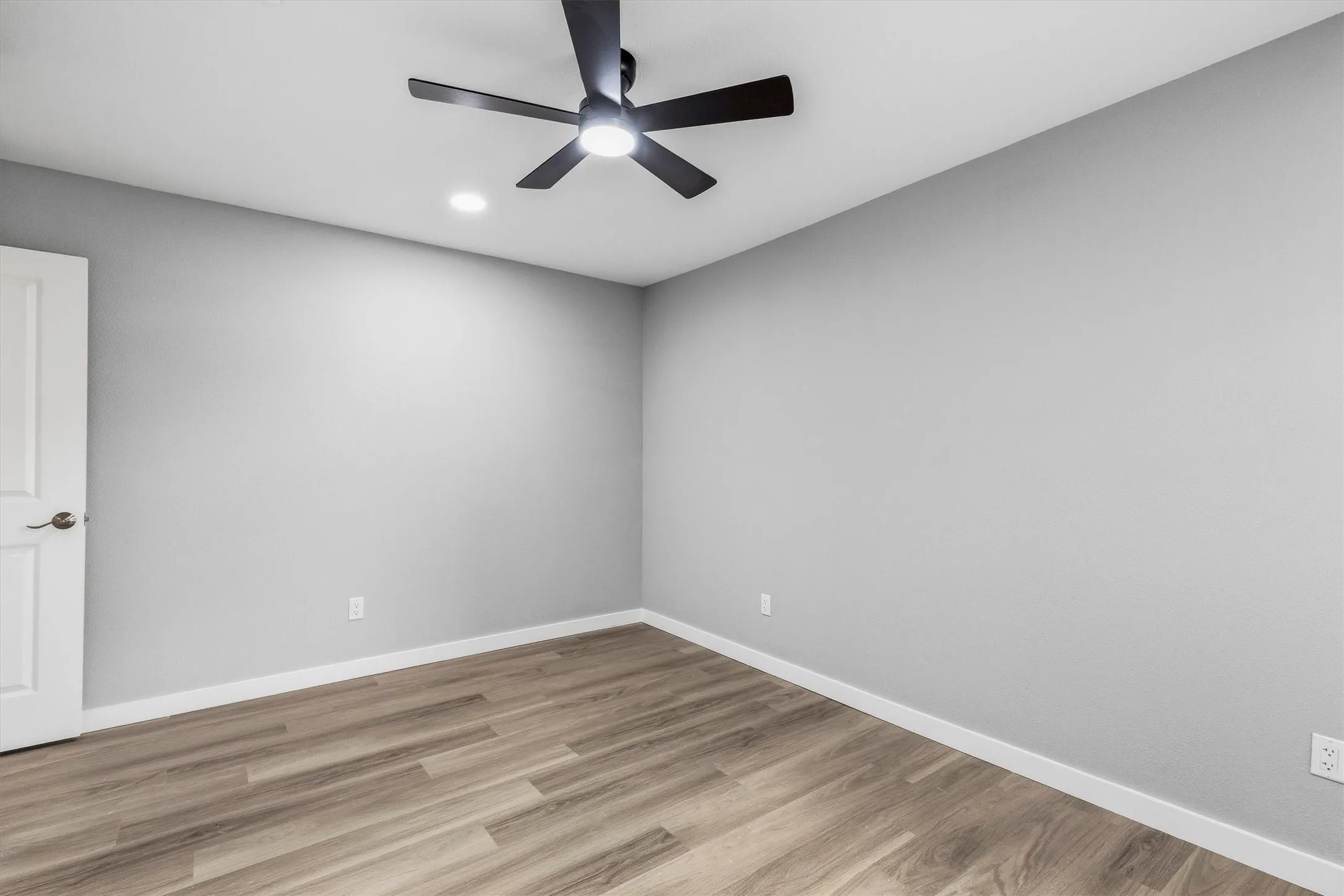 Wood-finish flooring with light gray walls, a white paneled door, and a black ceiling fan with integrated lighting