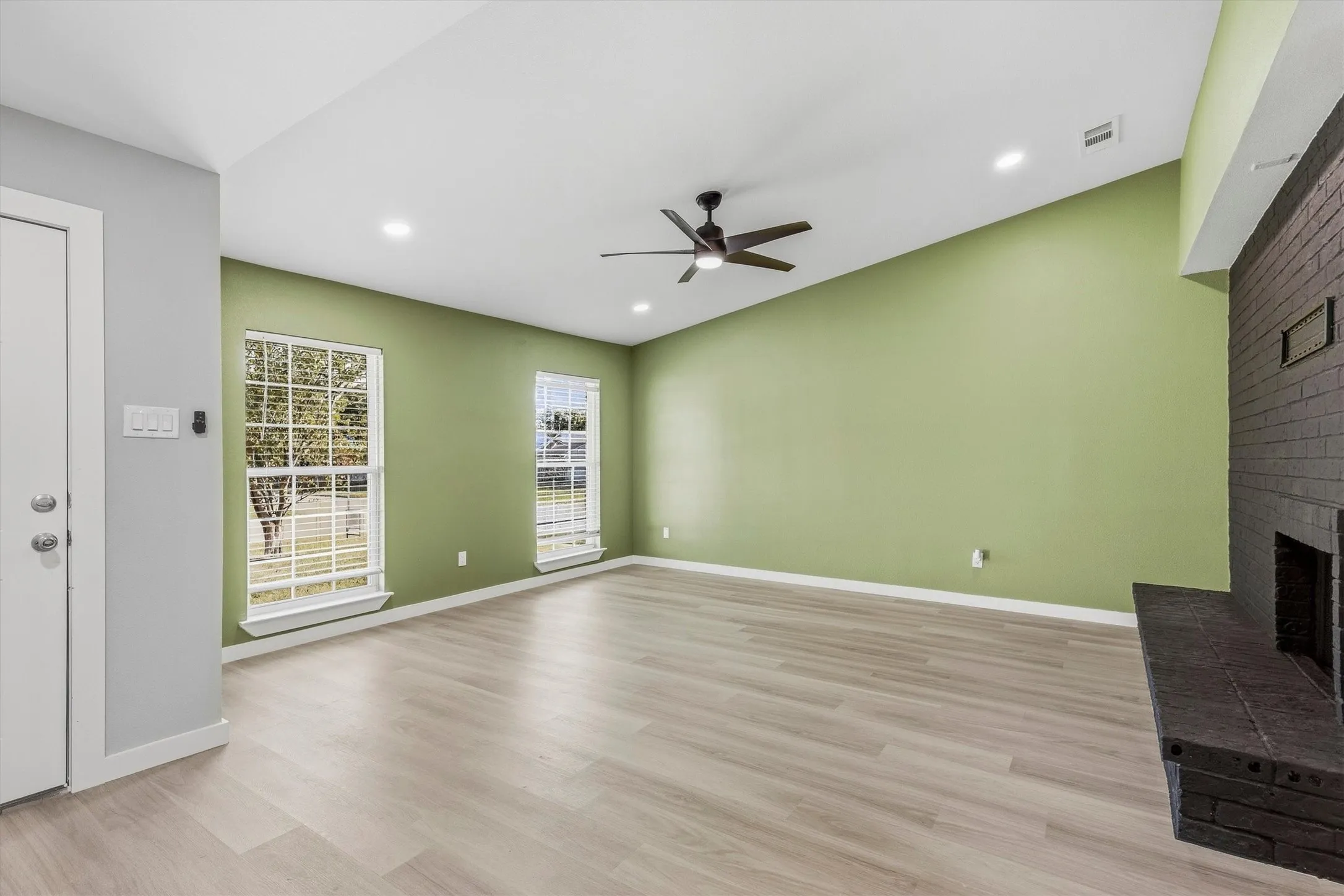Vaulted ceiling with a ceiling fan, recessed lighting, wood-finish flooring, two large windows, and a brick fireplace