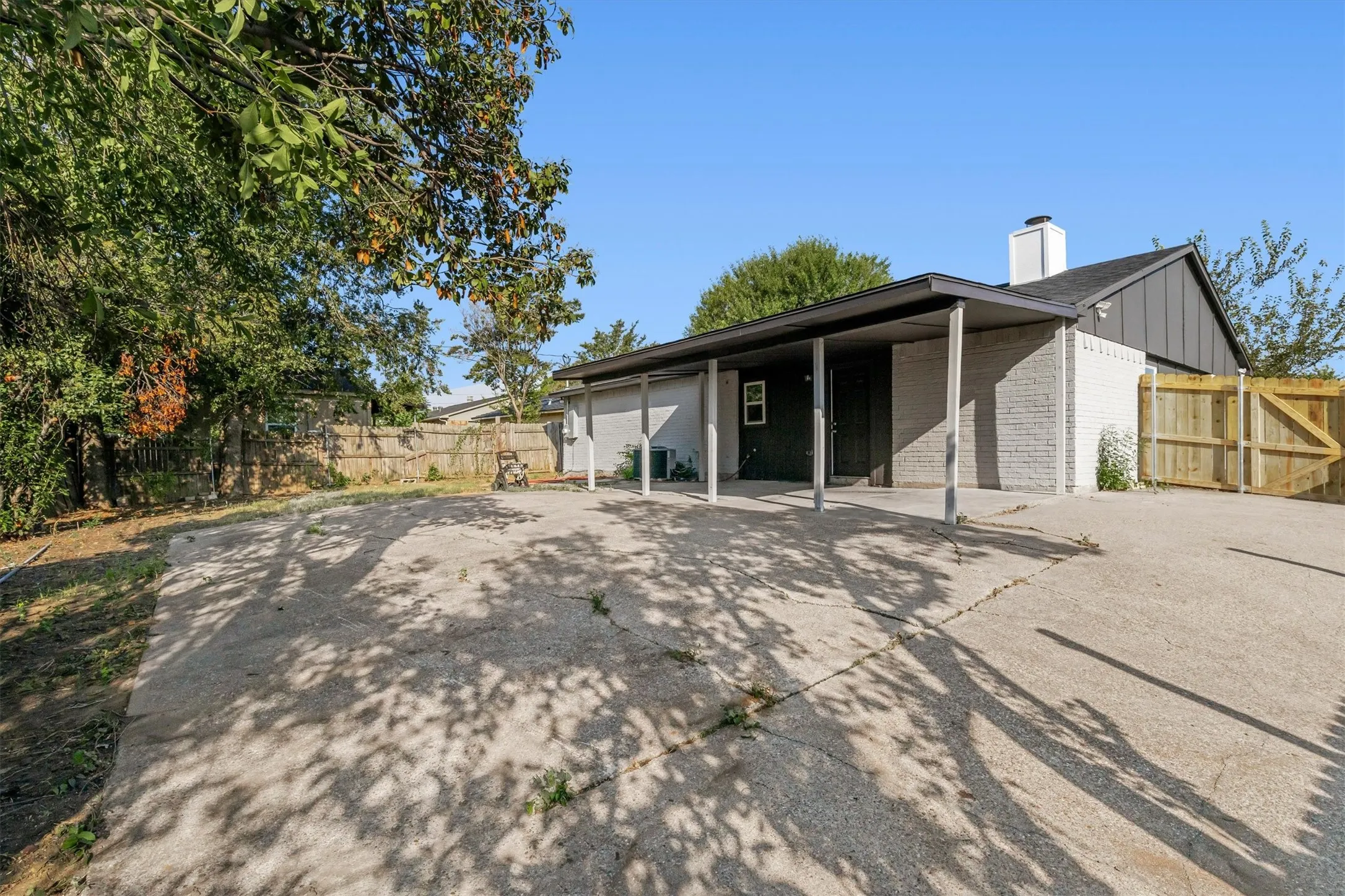 Concrete driveway with an attached carport, light-colored brick exterior, dark siding, and a wood-paneled fence