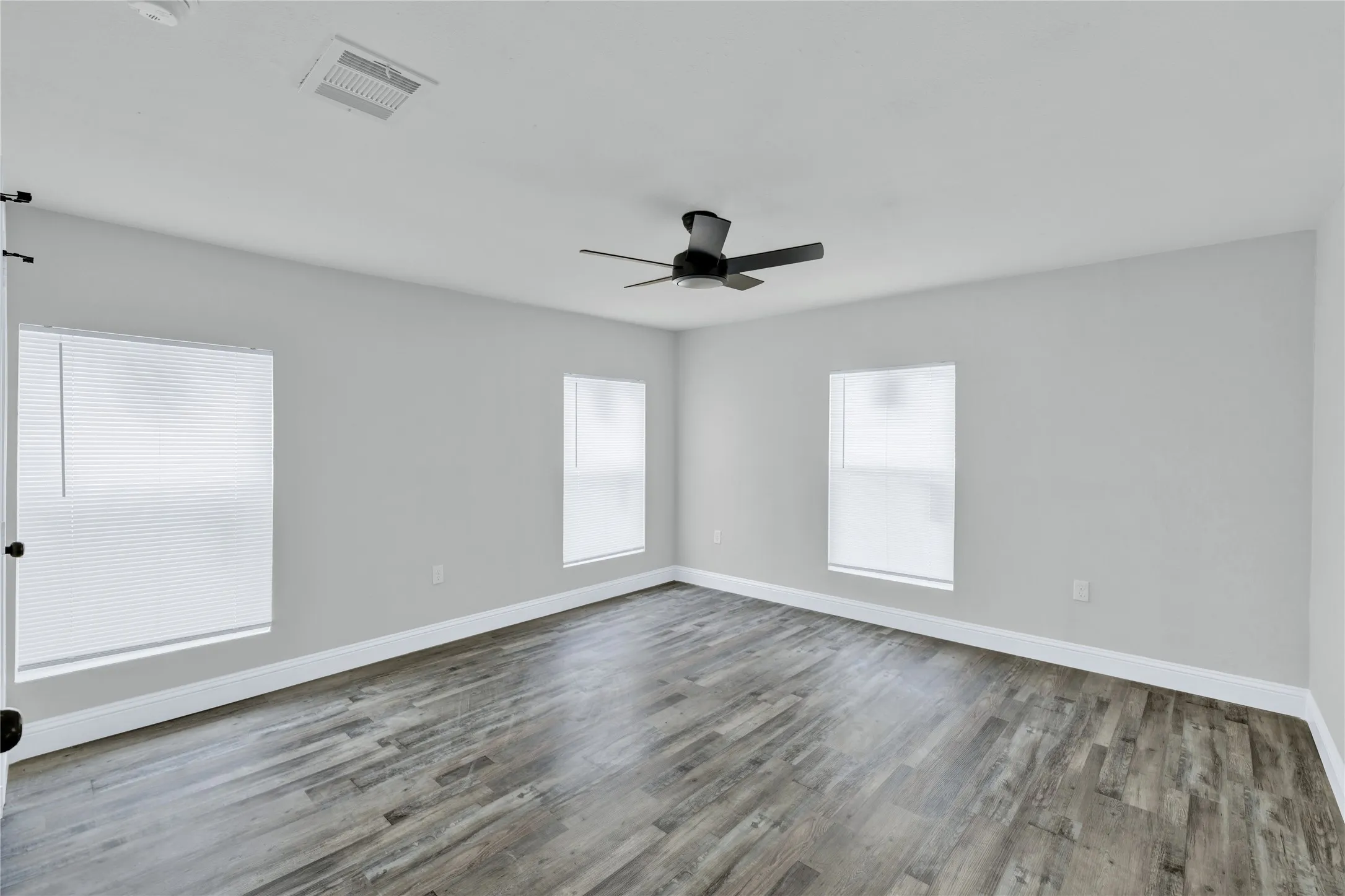 Spacious room featuring wood-finish flooring, light gray walls, multiple windows, and a ceiling fan