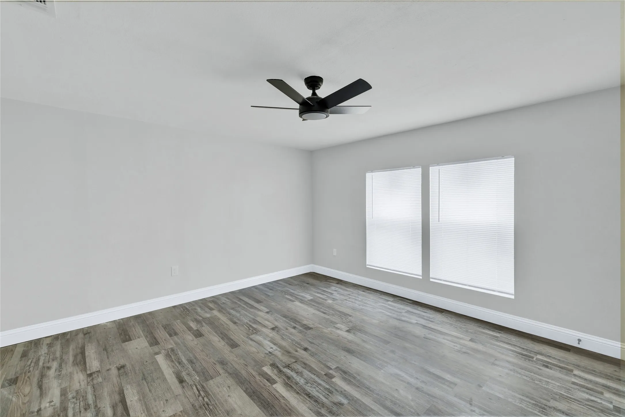Wood-finish flooring, white walls, two windows with blinds, and a ceiling fan
