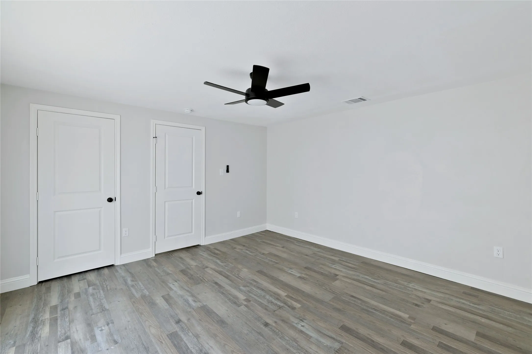 Wood-finish flooring, white walls, two paneled doors with black hardware, and a matte black ceiling fan with integrated lighting