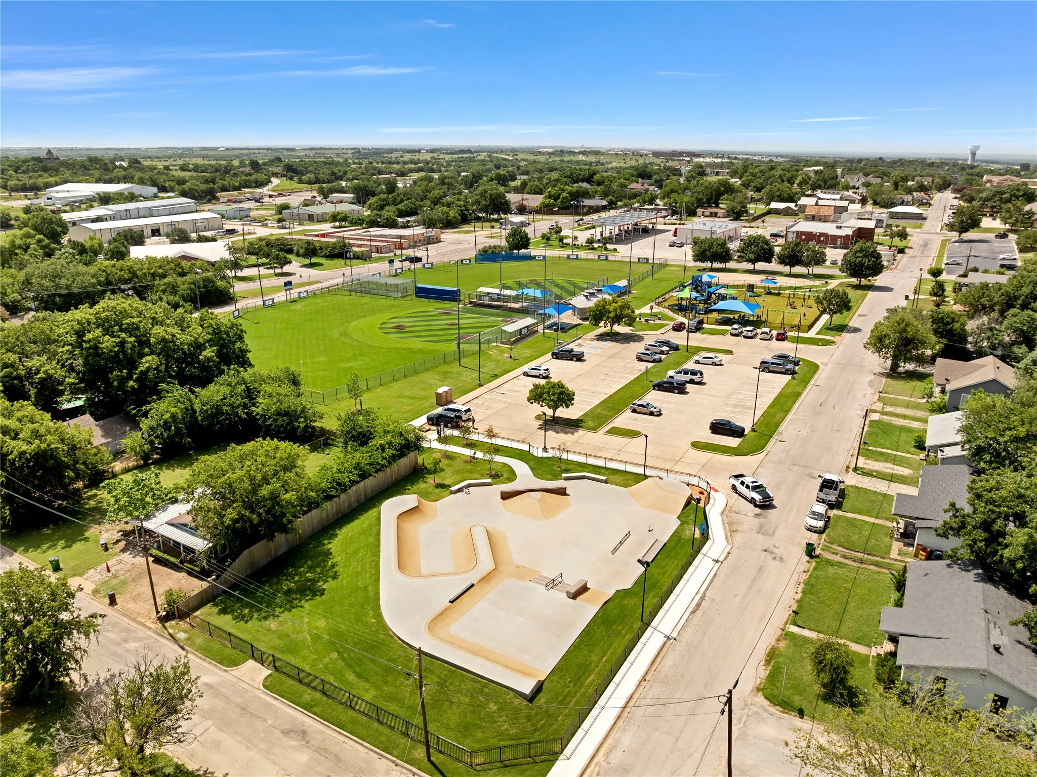 Open-air concrete skate park, adjacent green space, paved parking area, nearby baseball field, and recreational playground structures