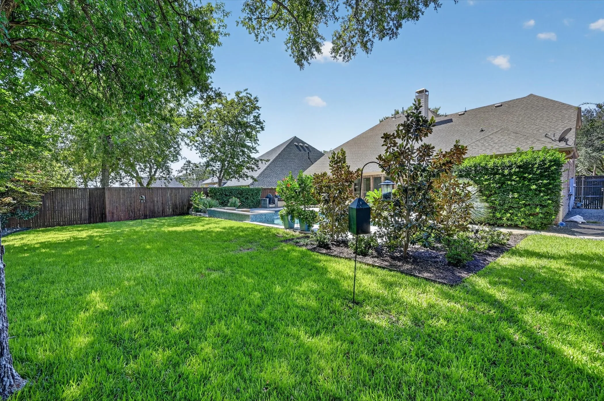 Expansive lawn area with mature trees, established landscaping beds, a wood privacy fence, and a visible swimming pool