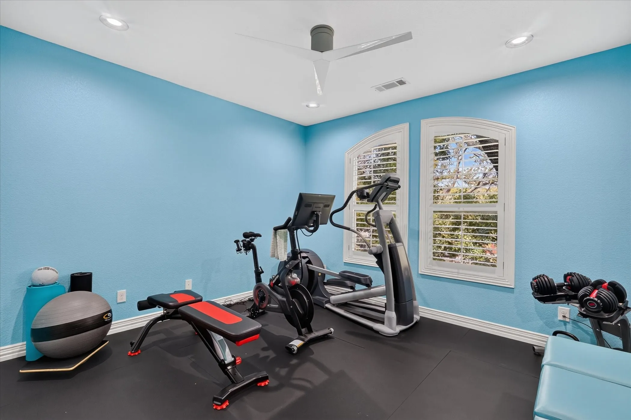 Recessed ceiling lighting, a modern ceiling fan, two arched windows with plantation shutters, and dark rubberized flooring