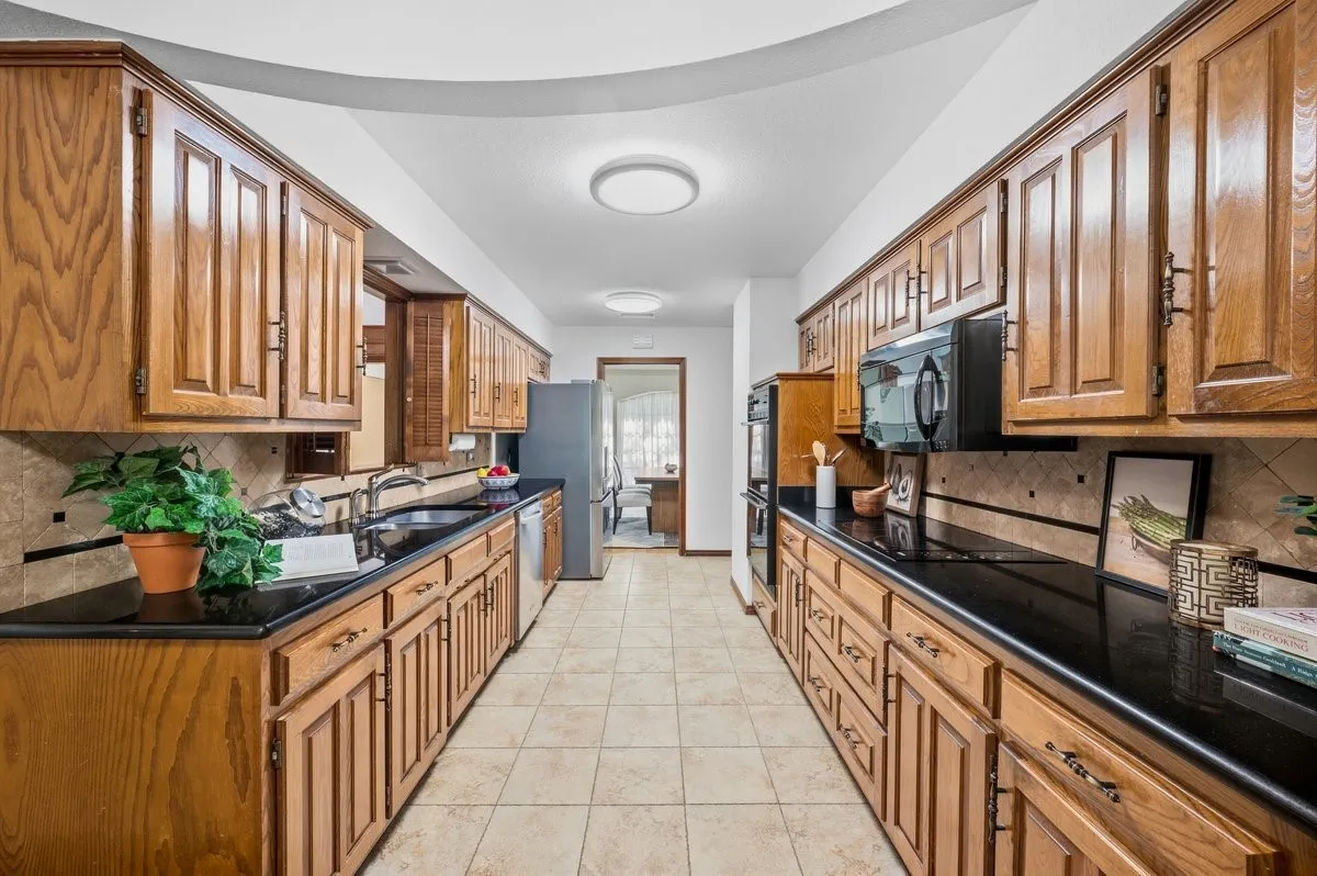 Galley kitchen featuring wood cabinetry, black countertops, tile backsplash, and stainless steel appliances
