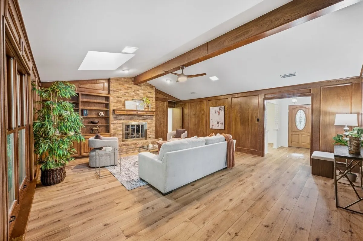 Spacious living area featuring wood-finish flooring, a brick fireplace, built-in shelving, wood paneled walls, and a vaulted ceiling with exposed beams