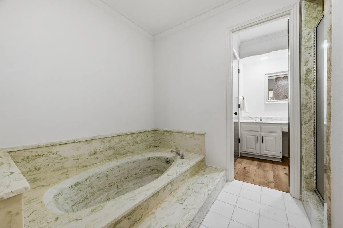Built-in bathtub with green-hued stone surround, white tiled flooring, and crown molding