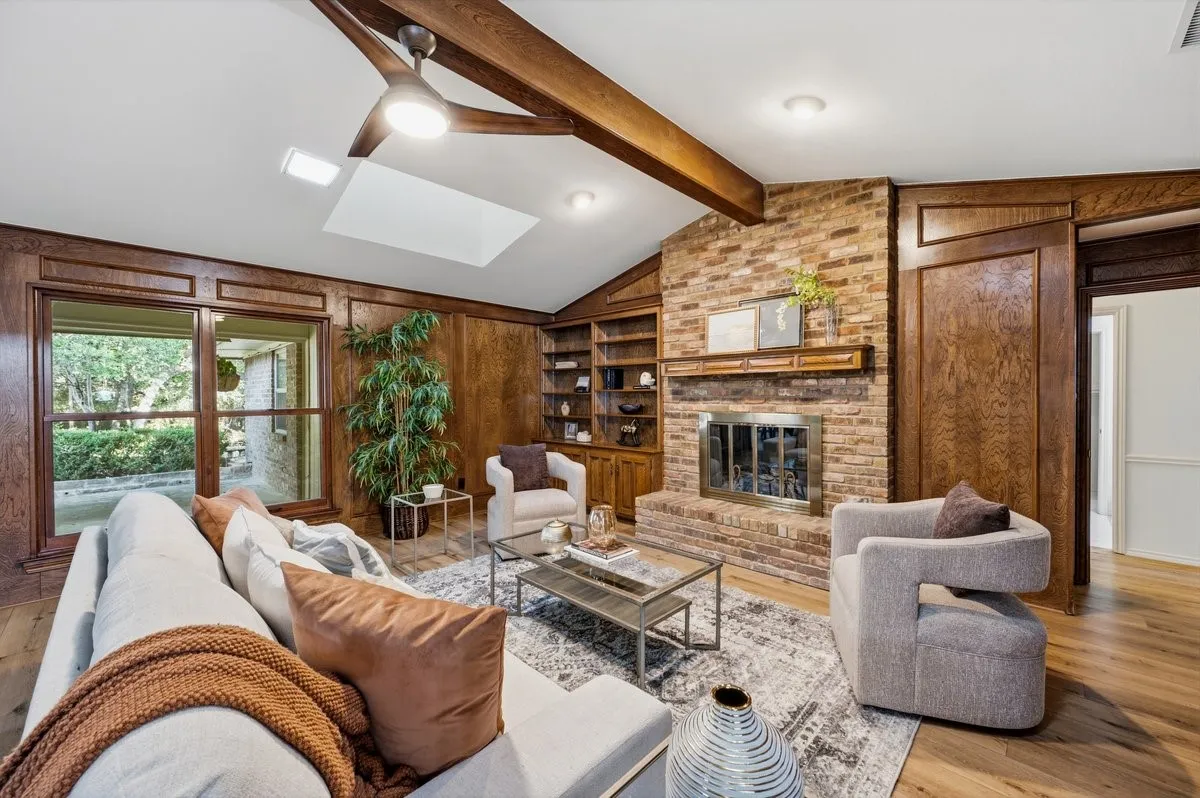 Vaulted ceiling with exposed wood beams, brick fireplace with a wood mantel, wood-finish flooring, built-in shelving, and large windows