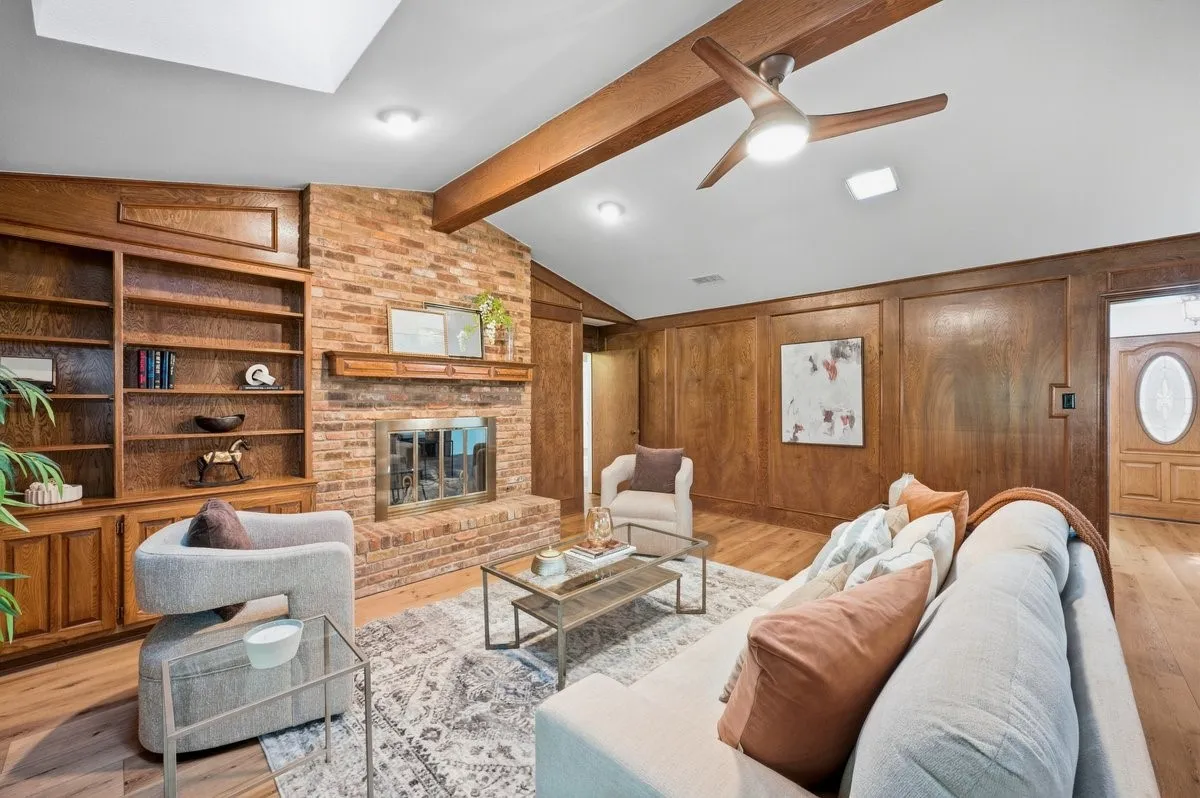 Vaulted ceiling with exposed wood beams, brick fireplace with wood mantel, built-in wooden shelving, wood-paneled walls, and wood-finish flooring