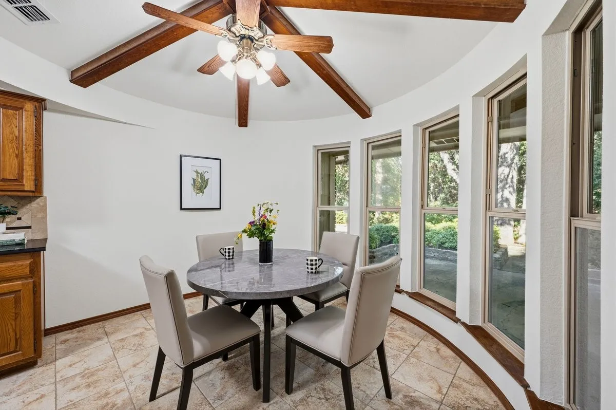 Kitchen area featuring wood-finish cabinetry with dark countertops, tiled flooring, wood-beamed ceiling with a ceiling fan, and a curved wall of multiple windows