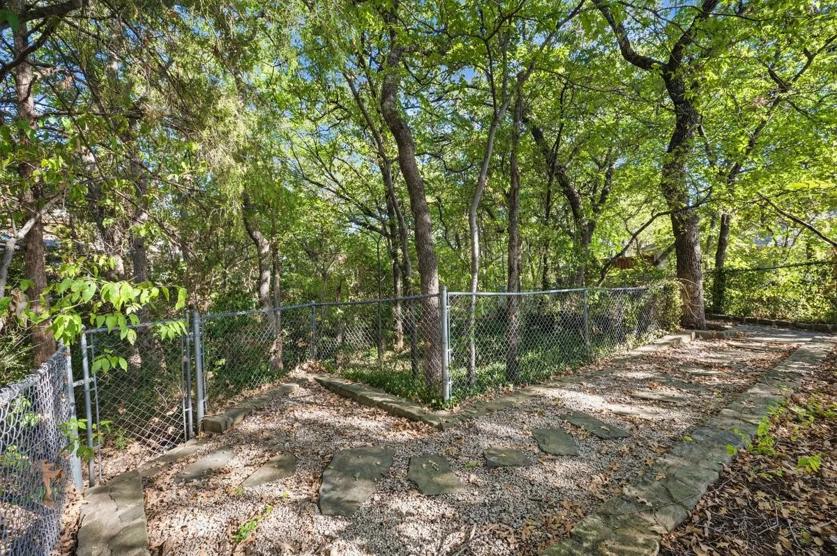 Wooded property featuring mature trees, a chain-link fence with gate, and a stone-edged path with stepping stones