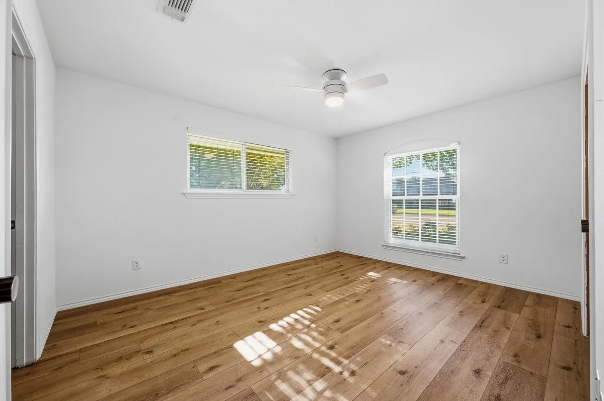 Spacious room featuring wood-finish flooring, two windows with blinds, a ceiling fan with integrated lighting, and neutral wall tones