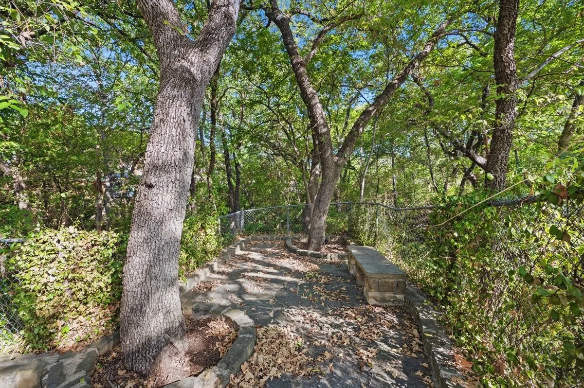 Stone pathway with integrated stone bench and mature trees, bordered by a chain-link fence and dense foliage